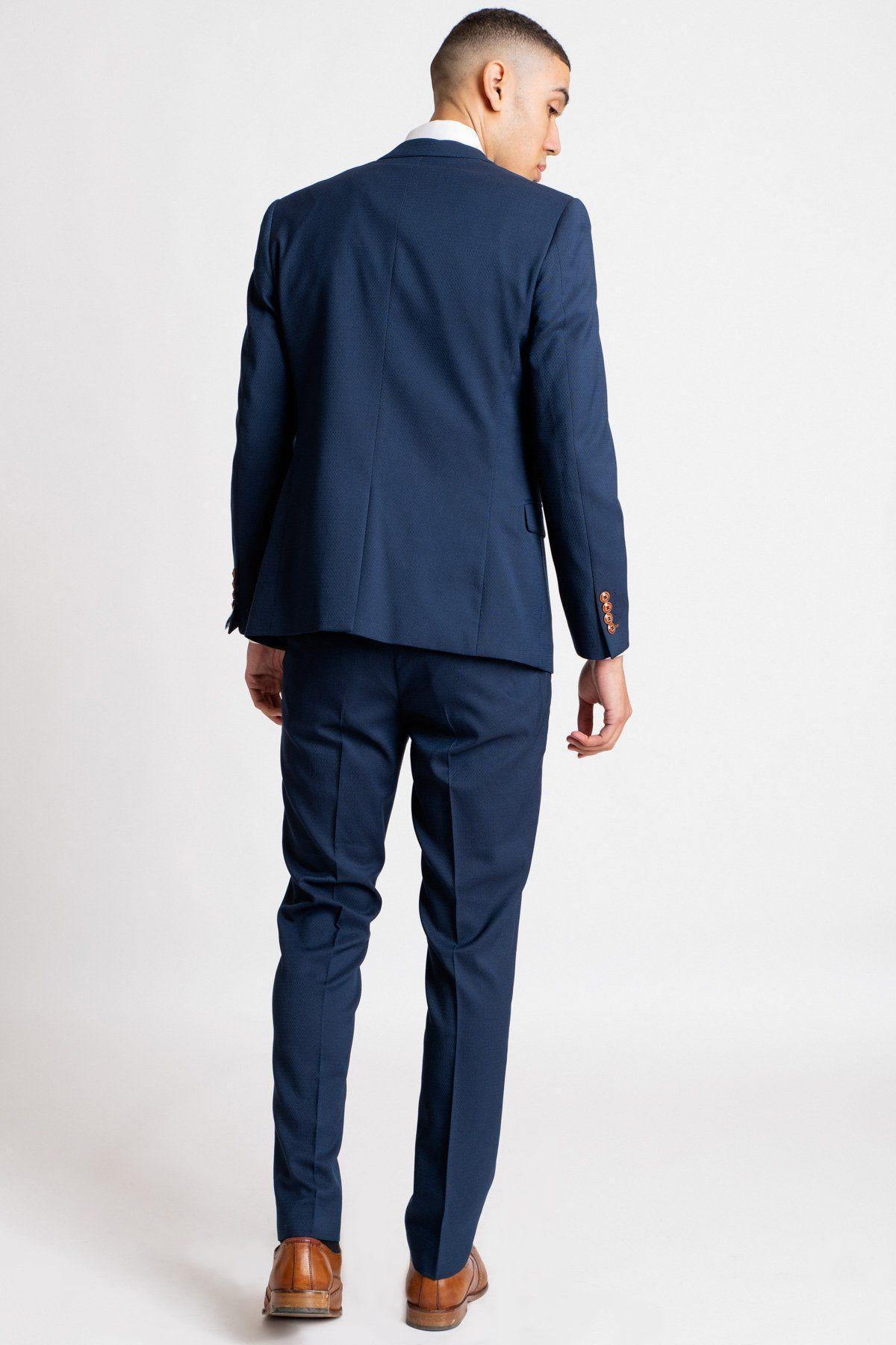 Owen Warner, 2022 runner-up of 'I'm A Celebrity Get Me Out Of Here', wears a marcdarcy Max Royal Blue Suit with brown shoes and a camouflage print pocket square, standing with his back to the camera against a white background.