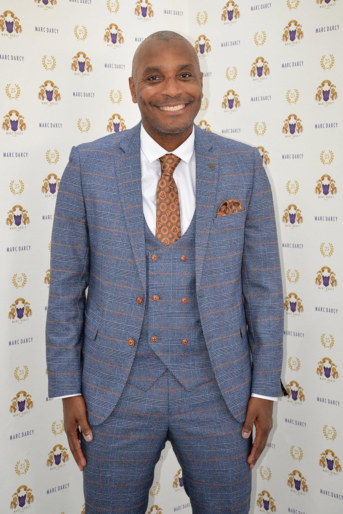 Former footballer Clinton Morrison wears the marcdarcy Jenson Sky Blue Check Suit, paired with a brown patterned tie and pocket square, smiling in front of a Marc Darcy step-and-repeat backdrop.