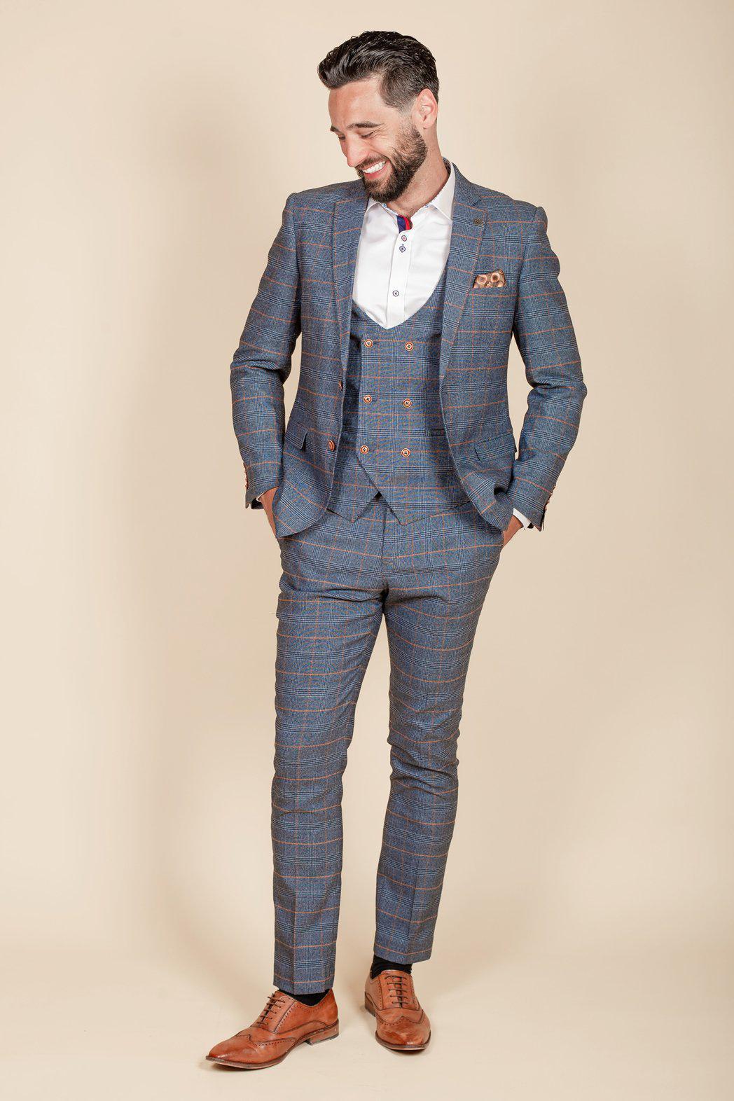 Clinton Morrison wears the marcdarcy Jenson Sky Blue Check Suit with a white shirt, brown dress shoes, and a patterned pocket square, smiling with hands in pockets against a beige background.