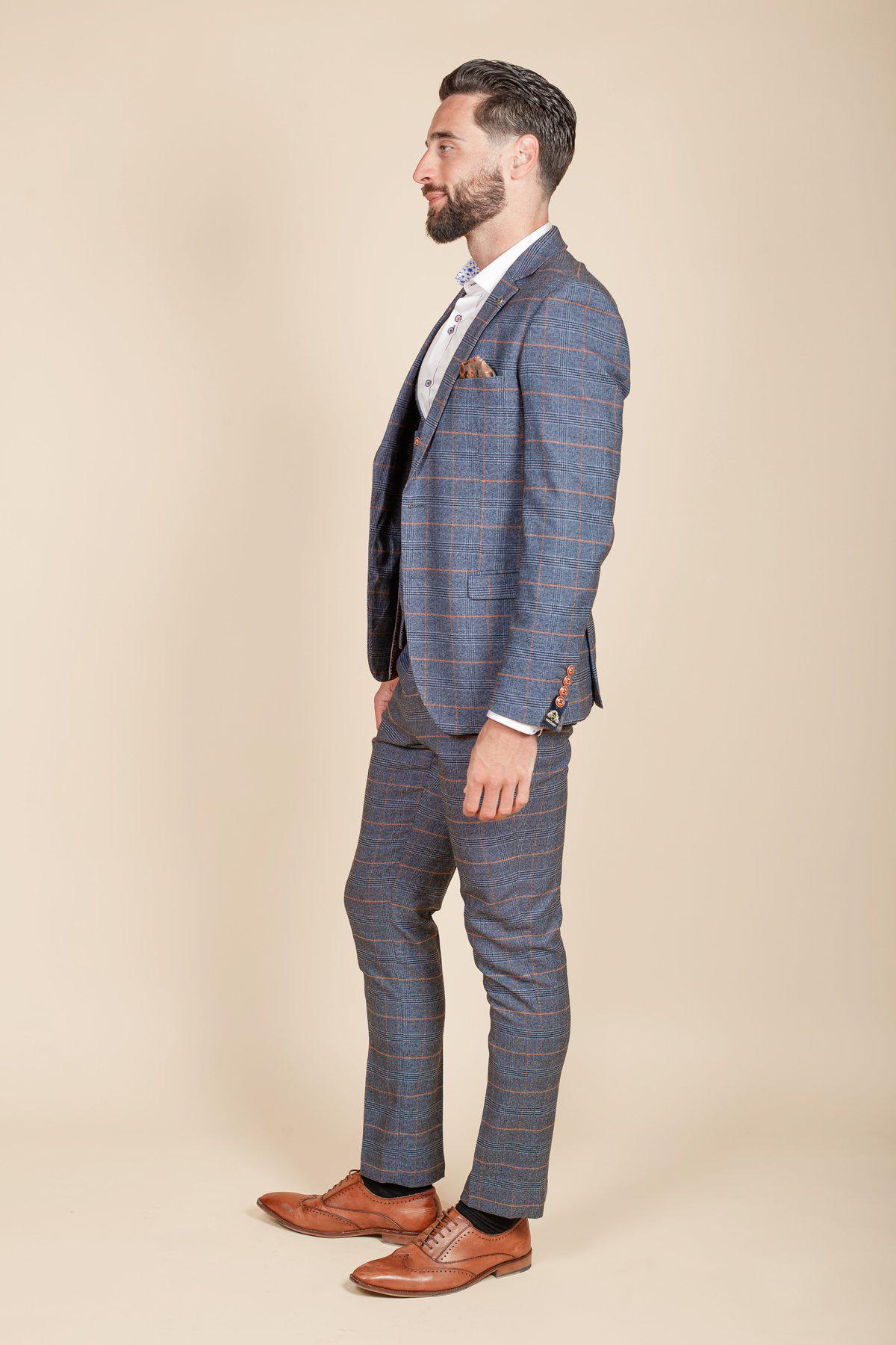 Former Footballer Clinton Morrison wears the marcdarcy Jenson Sky Blue Check three-piece suit with a white dress shirt, patterned pocket square, and brown dress shoes, standing in profile against a beige background.