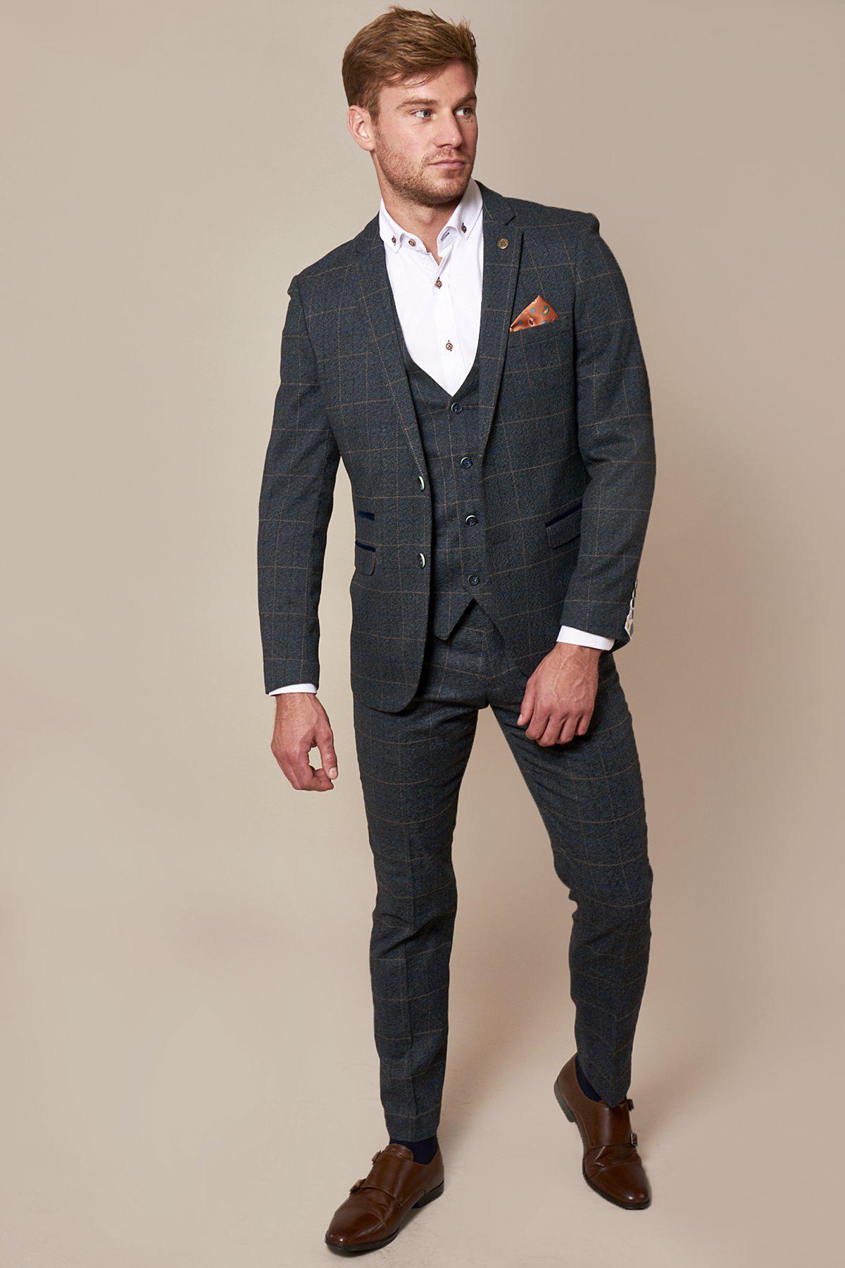 Weather presenter Alex Beresford wears the marcdarcy Scott Blue Tweed Check Suit: a vintage-inspired, dark blue check three-piece with navy velvet contrast, white shirt, brown belt and shoes, and an orange pocket square against a beige background.