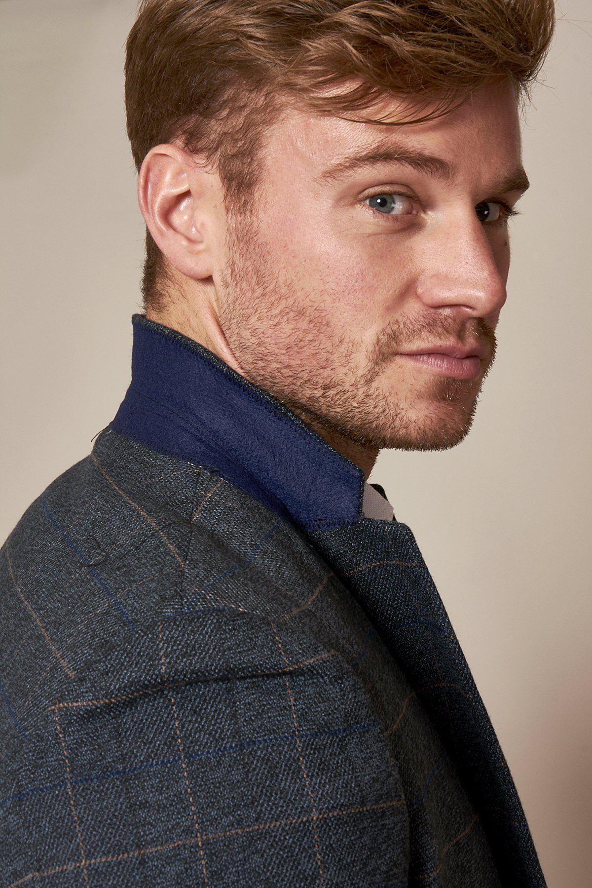 GMB Weather Presenter Alex Beresford, with short light brown hair and blue eyes, wears the marcdarcy Scott Blue Tweed Check Suit featuring a navy velvet contrast collar, looking over his shoulder against a neutral background.
