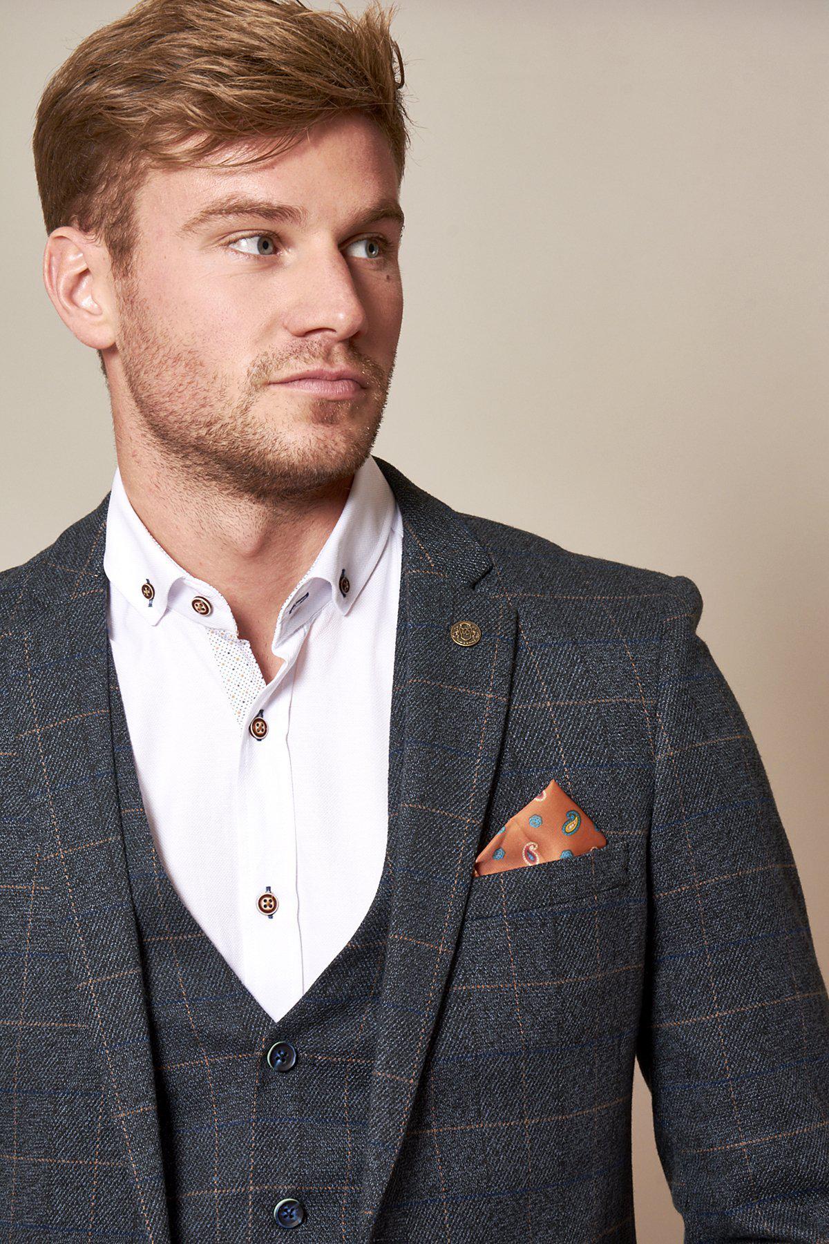 Alex Beresford wears the marcdarcy Scott Blue Tweed Check Suit—vintage-inspired with subtle navy velvet contrast—over a white shirt and patterned orange pocket square, gazing left with a neutral expression.