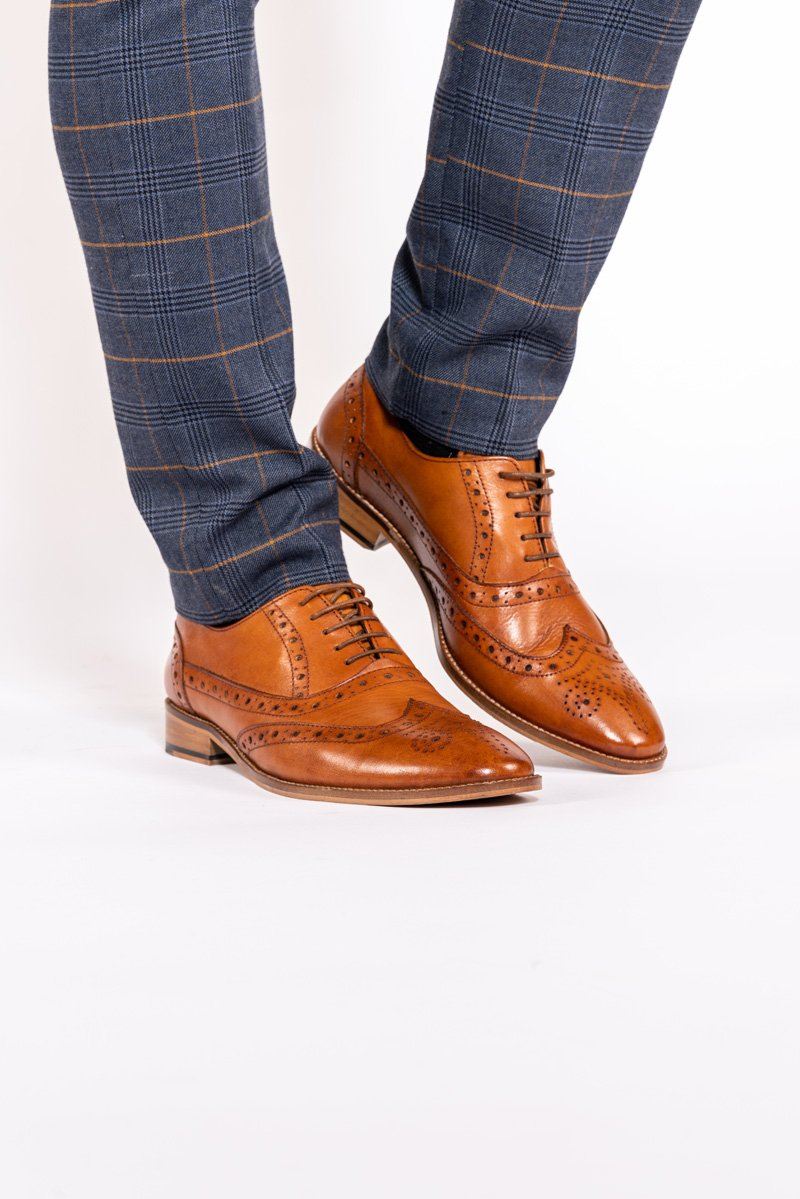 A person stands on a white background wearing slim-fit blue plaid trousers and Marc Darcys GRAYSON Tan Leather Wingtip Brogue Shoes featuring handcrafted detailing.