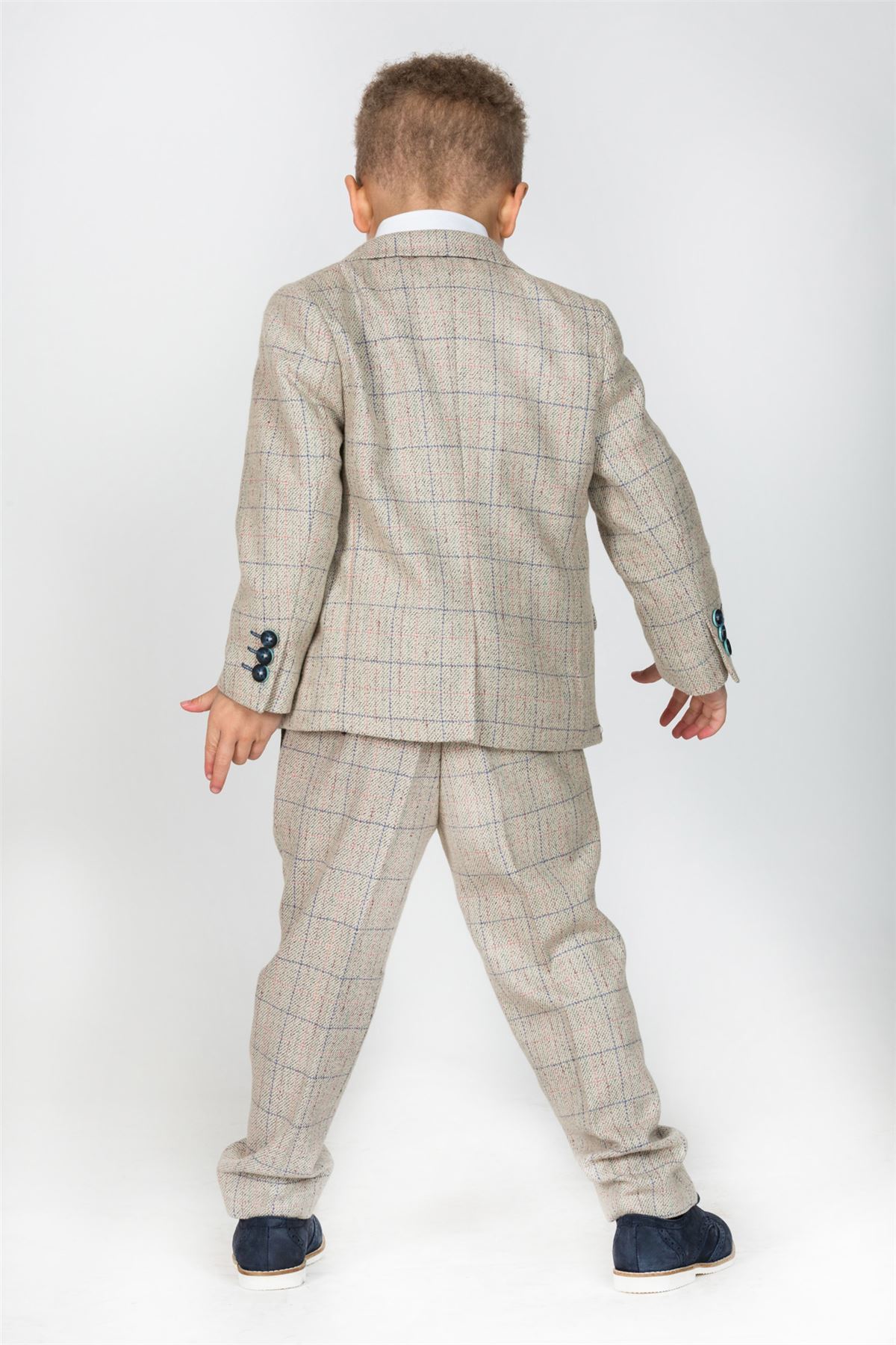 A young child with curly hair, seen from behind, wears the marcdarcy HARDING Childrens Cream Tweed Check Three Piece Suit, featuring matching trousers and jacket with luxury paisley lining, a white shirt, and dark shoes.