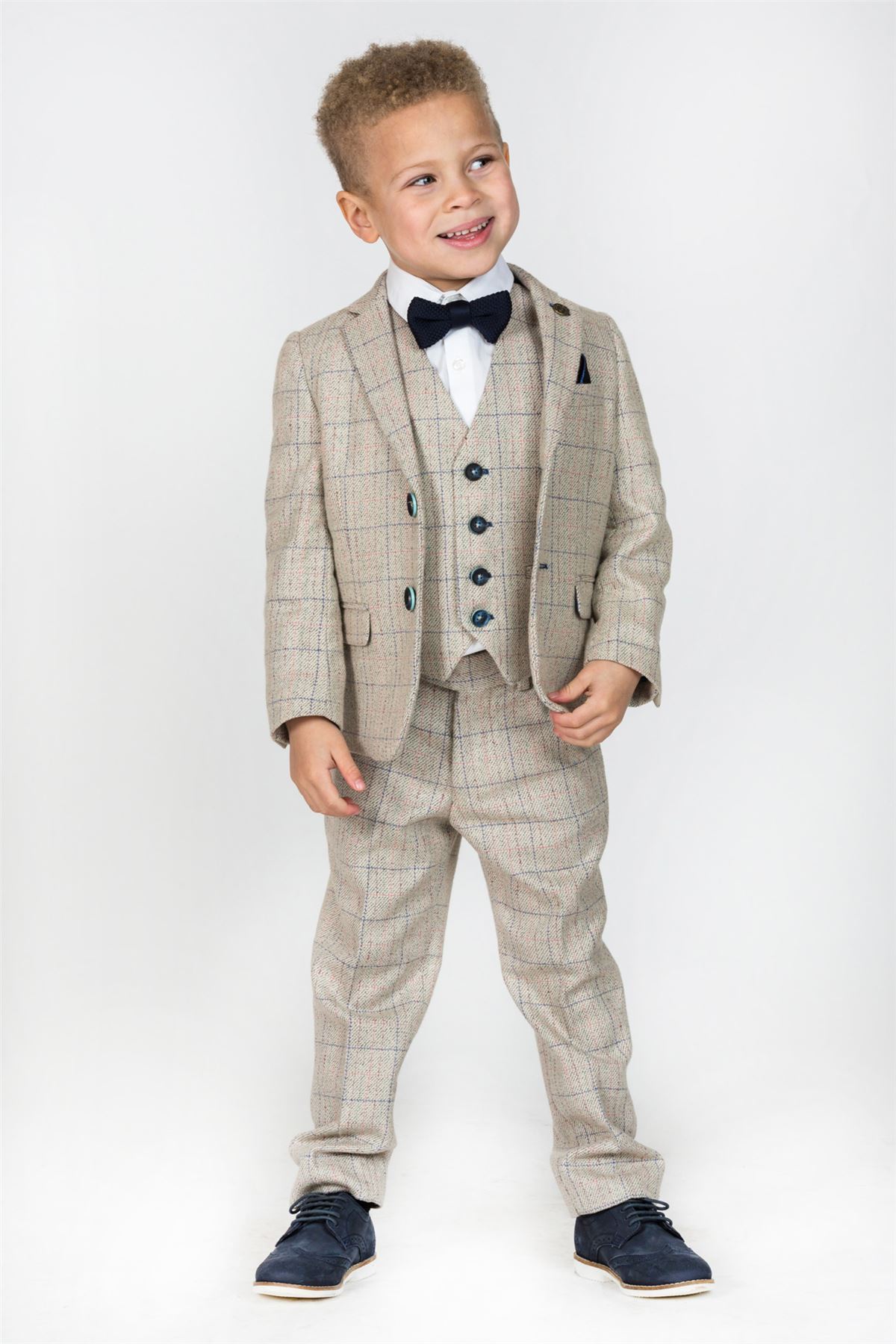 A young boy smiles while wearing the marcdarcy HARDING Childrens Cream Tweed Check Three Piece Suit, featuring a cream tweed check jacket, white shirt, navy bow tie, and dark shoes against a plain white background.