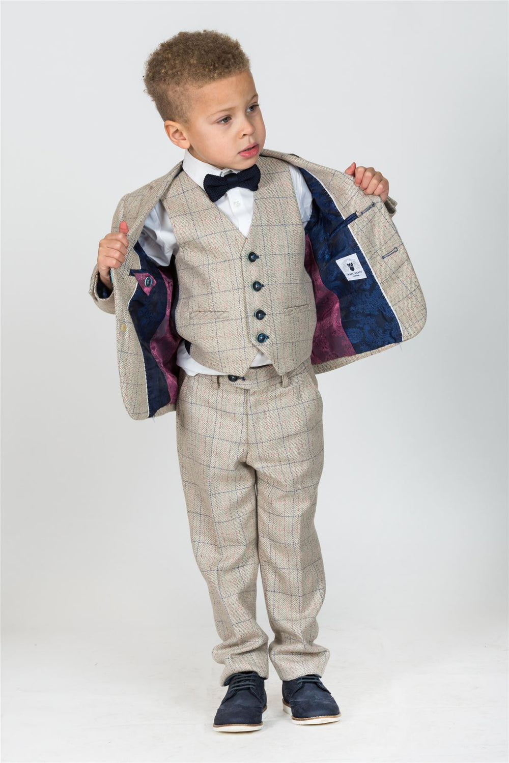 A young boy models the marcdarcy HARDING Childrens Cream Tweed Check Three Piece Suit, featuring a white shirt, navy bowtie, waistcoat, dress pants, navy shoes, and displaying the patterned blazer lining.