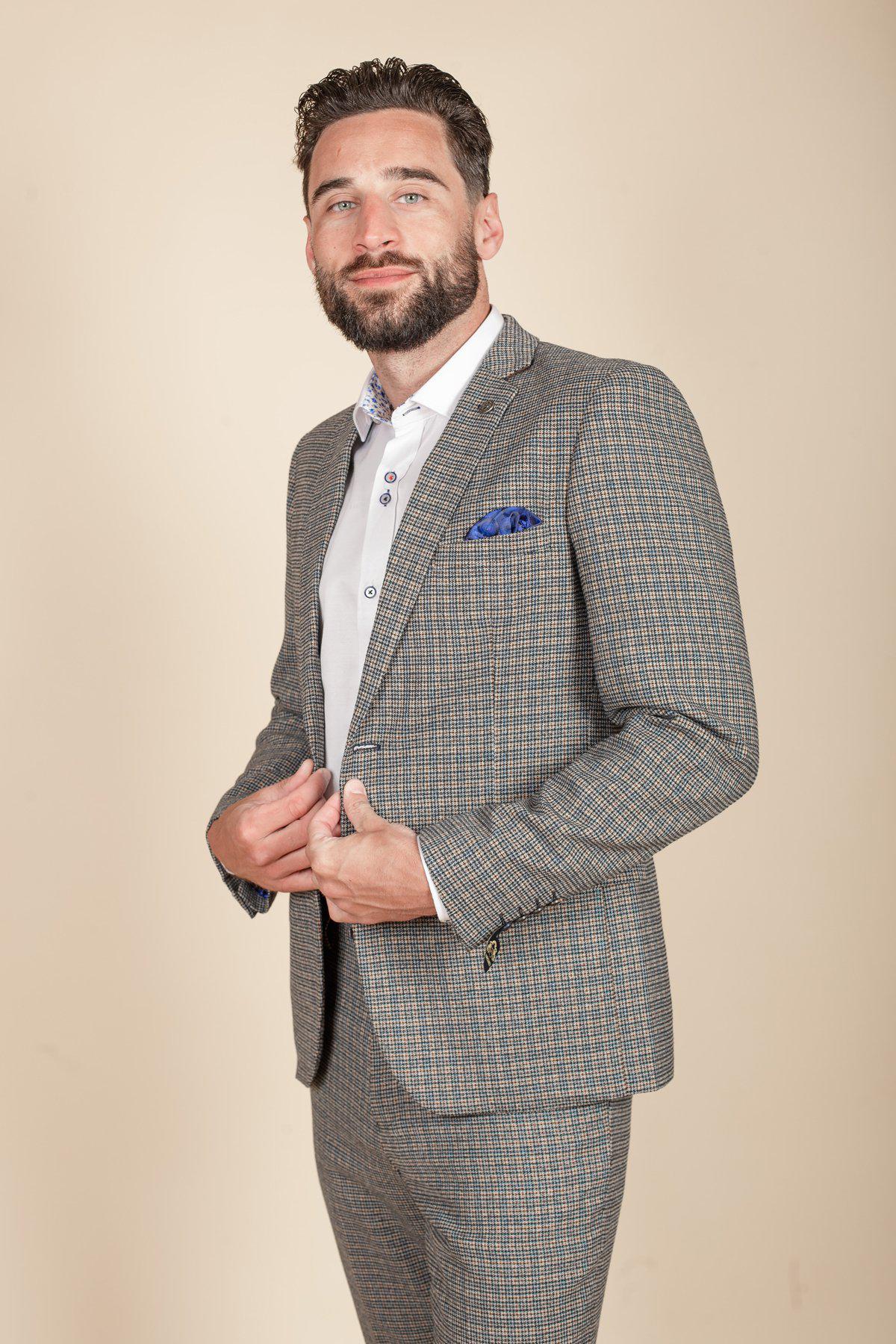 A man with short dark hair and a beard wears the marcdarcy HARDWICK Navy Tan Tweed Check Blazer over a white shirt with a blue pocket square, standing against a plain beige background while holding his jacket.