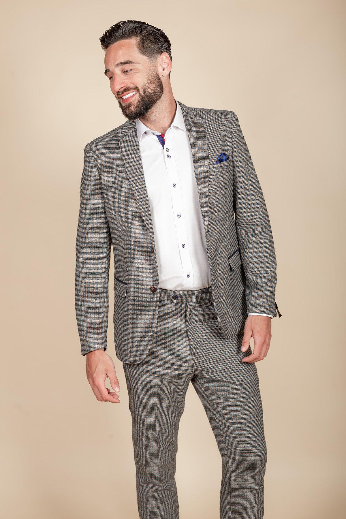 Bearded man smiling and looking to the side, wearing the marcdarcy HARDWICK - Navy Tan Tweed Check Blazer over a white dress shirt with a blue pocket square, standing against a plain beige background.