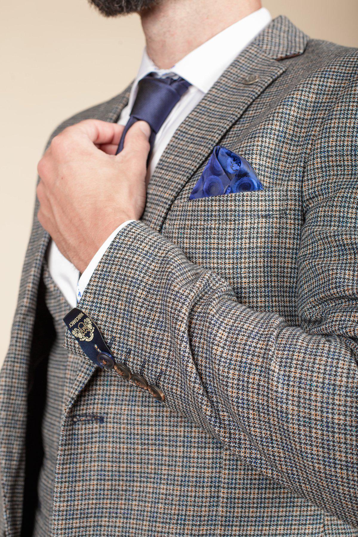 A man adjusts his tie while wearing a marcdarcy HARDWICK Navy Tan Tweed Check Blazer, paired with a white dress shirt, blue patterned tie, and blue pocket square. The image highlights his torso and hand, not showing his full face.