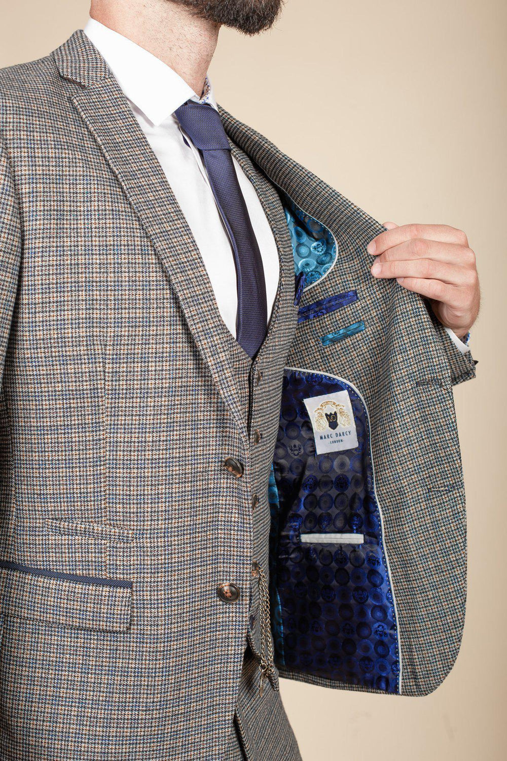 A man in the marcdarcy HARDWICK Navy Tan Tweed Check Blazer holds open his jacket to show its blue patterned satin lining and inner label; his plaid suit and navy tie enhance his distinguished style, though his face is partly out of frame.