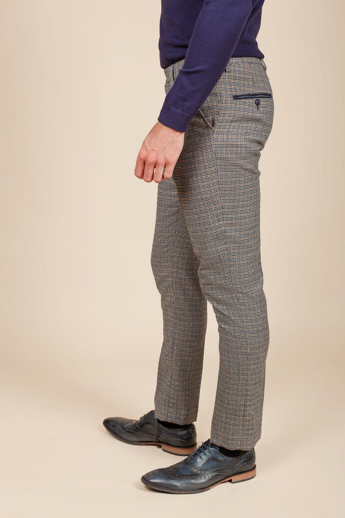 A person stands sideways, showing only their lower half, wearing marcdarcy HARDWICK Navy Tan Tweed Check Trousers in a slim fit with black dress shoes against a beige background.
