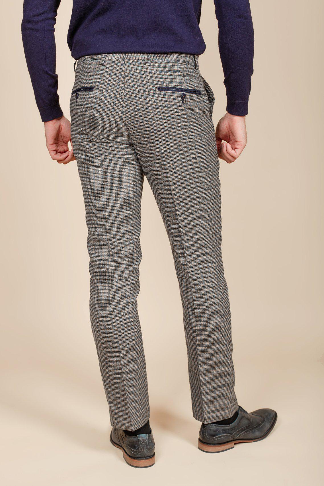 A man stands facing away, wearing marcdarcy HARDWICK - Navy Tan Tweed Check Trousers, a navy sweater, and dark dress shoes against a beige background. His hands rest by his sides.