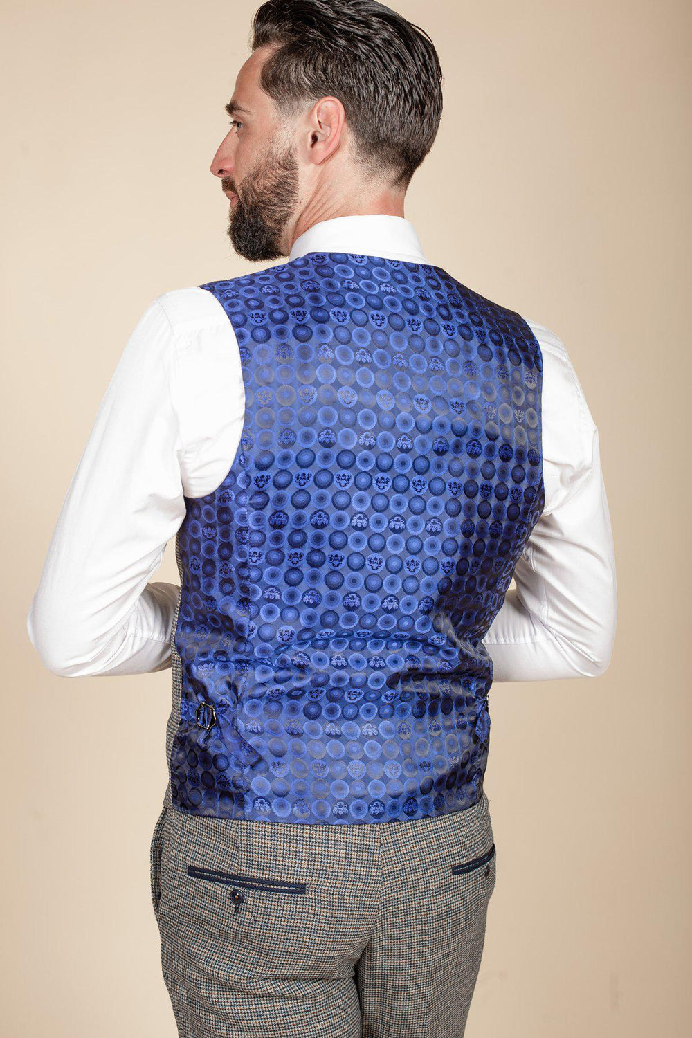 A man with dark hair and a beard is shown from the back, wearing a white dress shirt and the marcdarcy HARDWICK Navy Tan Tweed Check Waistcoat. He pairs it with gray heritage trousers. The background is beige.