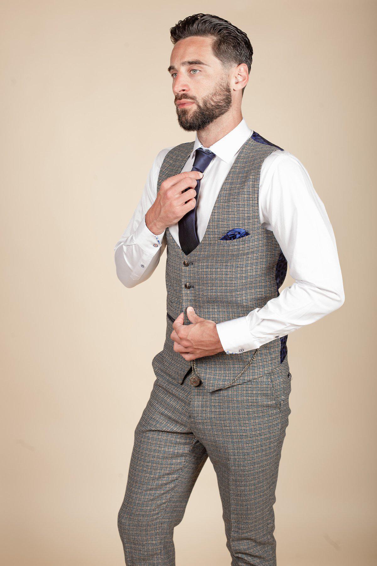 A man with short dark hair and a beard adjusts a navy tie while wearing marcdarcy's HARDWICK Navy Tan Tweed Check Waistcoat, grey heritage trousers, a white shirt, and blue pocket square against a plain beige background.