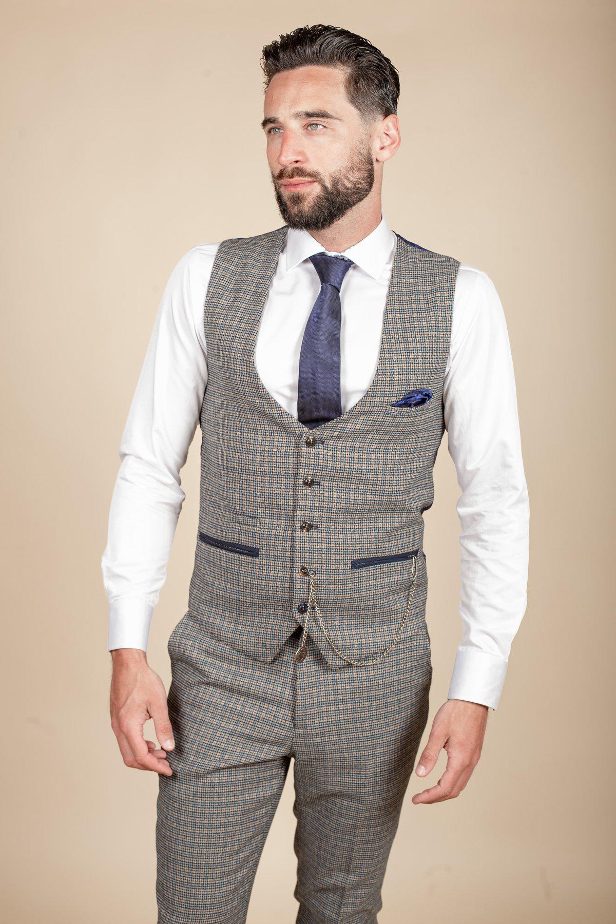 A man with dark hair and a beard wears the marcdarcy HARDWICK Navy Tan Tweed Check Waistcoat as part of a gray three-piece suit, paired with a white shirt, navy tie, and pocket square. He stands against a beige background, looking slightly to the side.