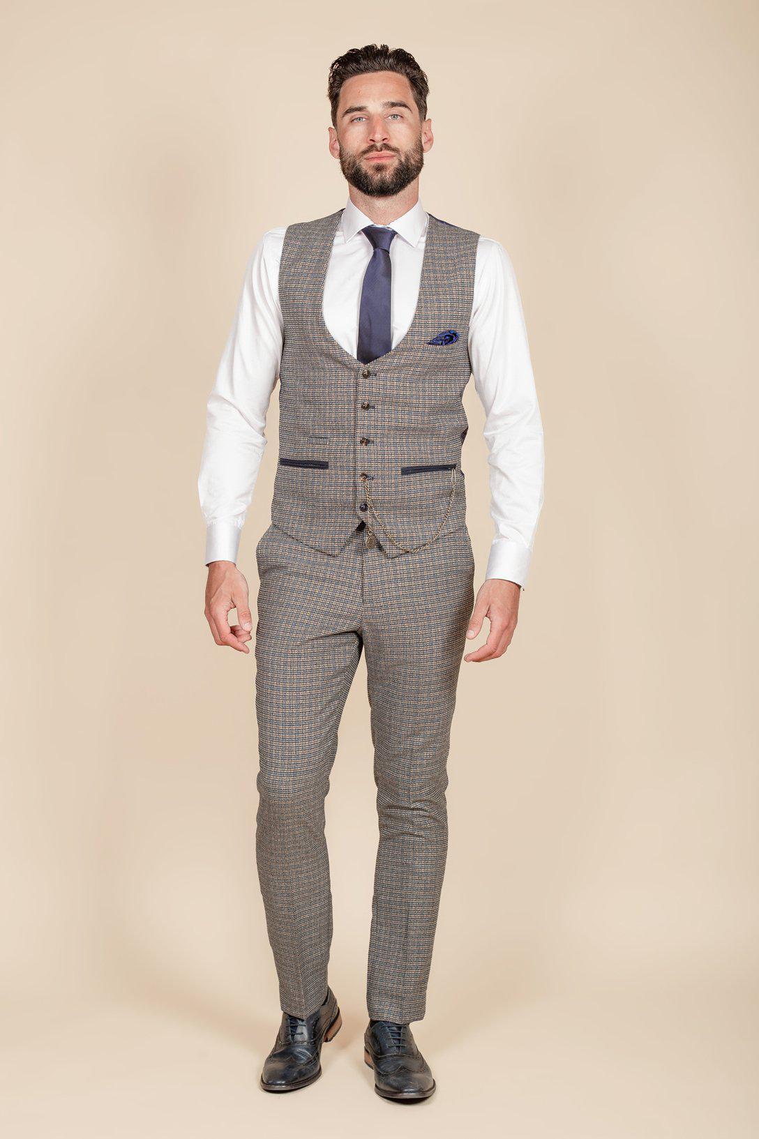 A man faces forward wearing the marcdarcy HARDWICK navy tan tweed check waistcoat, paired with heritage style trousers, a white shirt, navy tie, pocket square, and black dress shoes against a plain beige background.