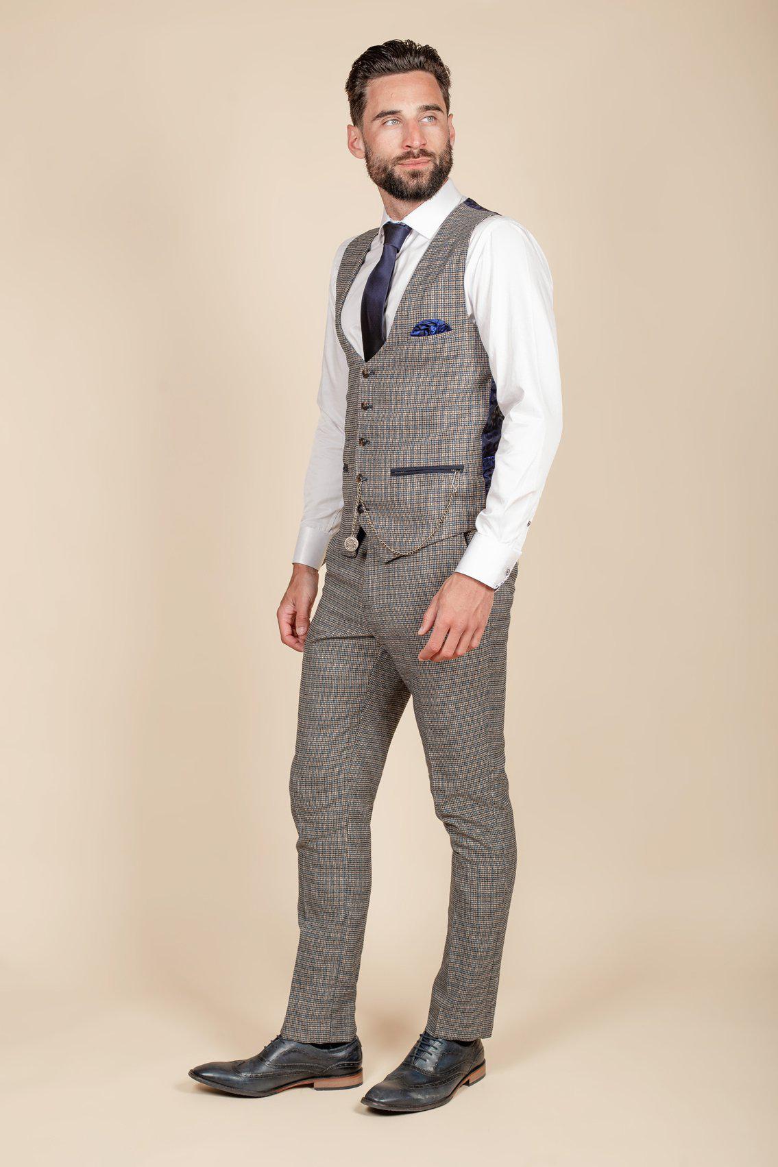 Man with a beard wearing a marcdarcy HARDWICK Navy Tan Tweed Check Waistcoat, grey checkered three-piece suit, white shirt, navy tie, and black dress shoes, standing and looking to the side against a beige background.