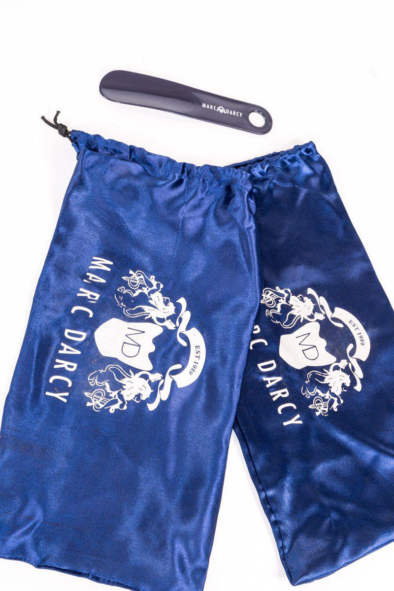A dark blue Marc Darcy shoehorn rests above two flat, hand-stitched Marc Darcy drawstring bags with white logos, styled alongside the HARRIS black leather double strap monk shoes.