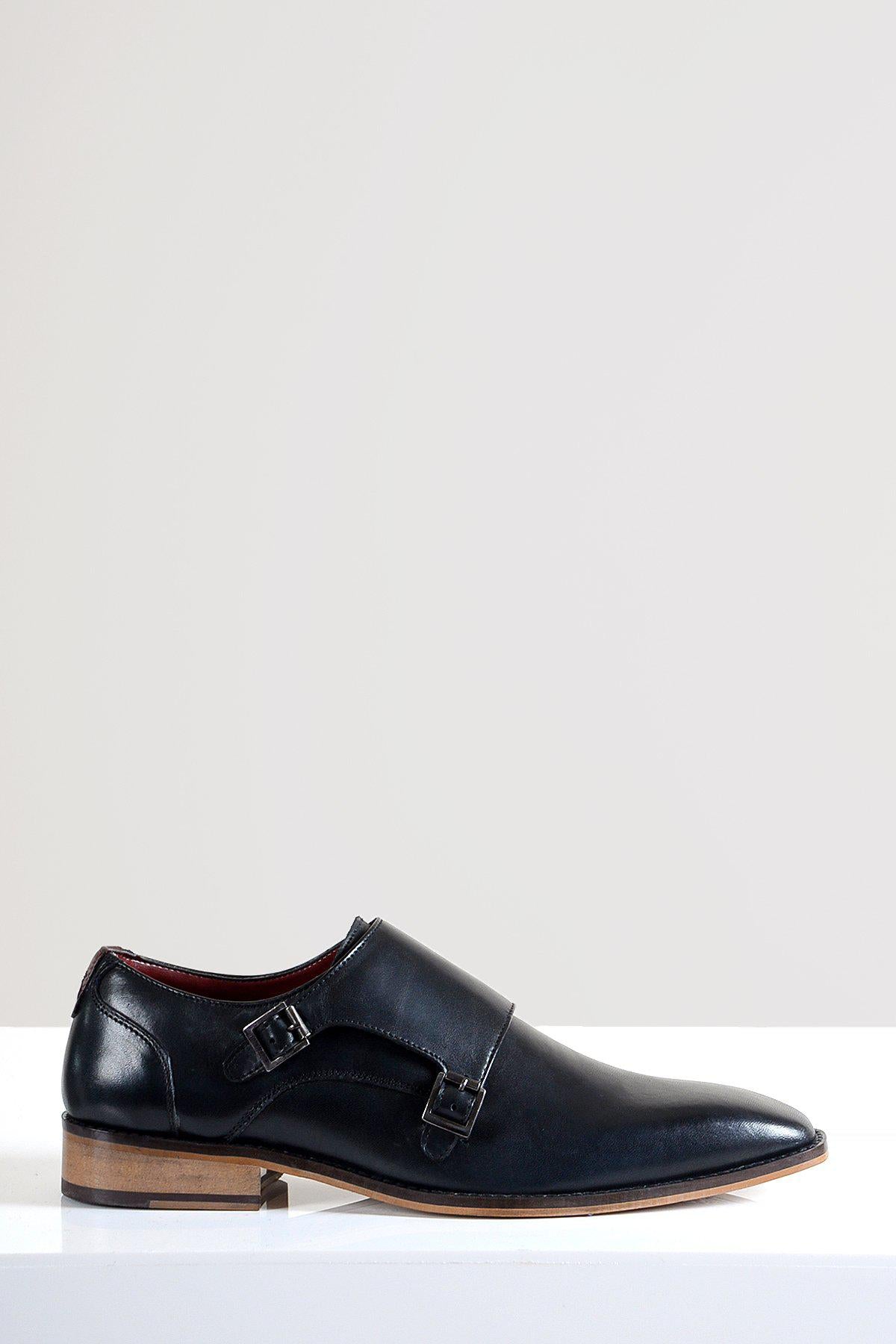 The Marc Darcy HARRIS Black Leather Double Strap Monk Shoe, crafted from full grain leather with a light brown sole, is displayed on a white surface against a plain light gray background.