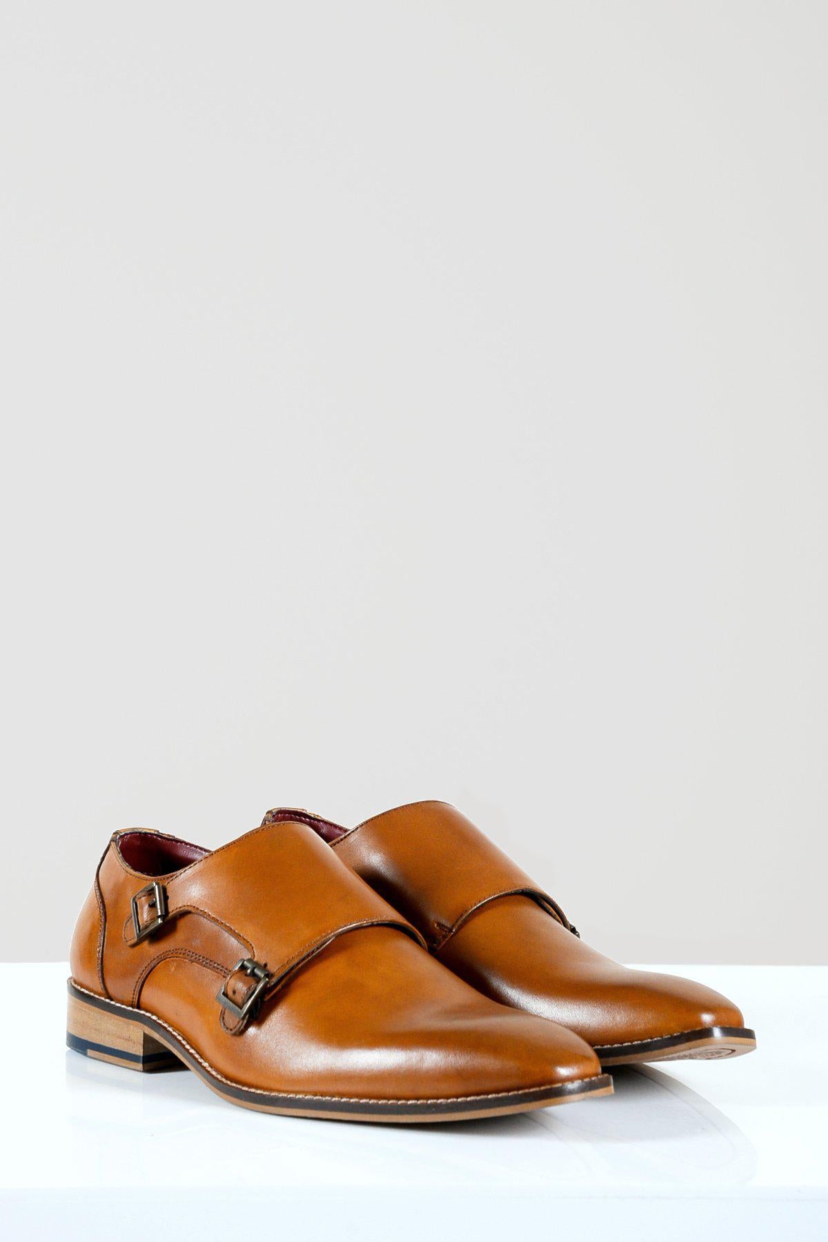 The Marc Darcy HARRIS Tan Leather Double Strap Monk Shoe features gunmetal buckles, pointed toes, and a wooden heel with navy accent, displayed on a white surface against a light grey background.