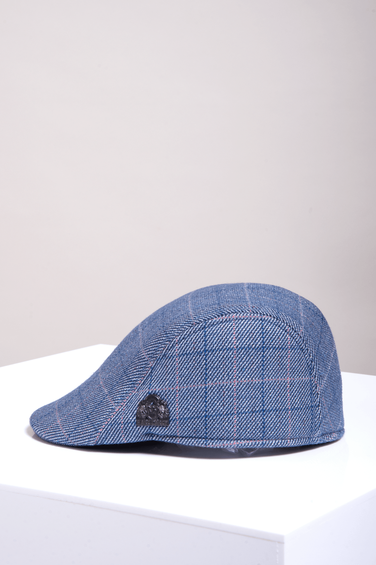 The HARRY Blue Tweed Check Flat Cap by Accessory, featuring blue plaid with red pinstripe detail and a black emblem, is styled in Marc Darcy fashion and placed on a white surface against a light background.