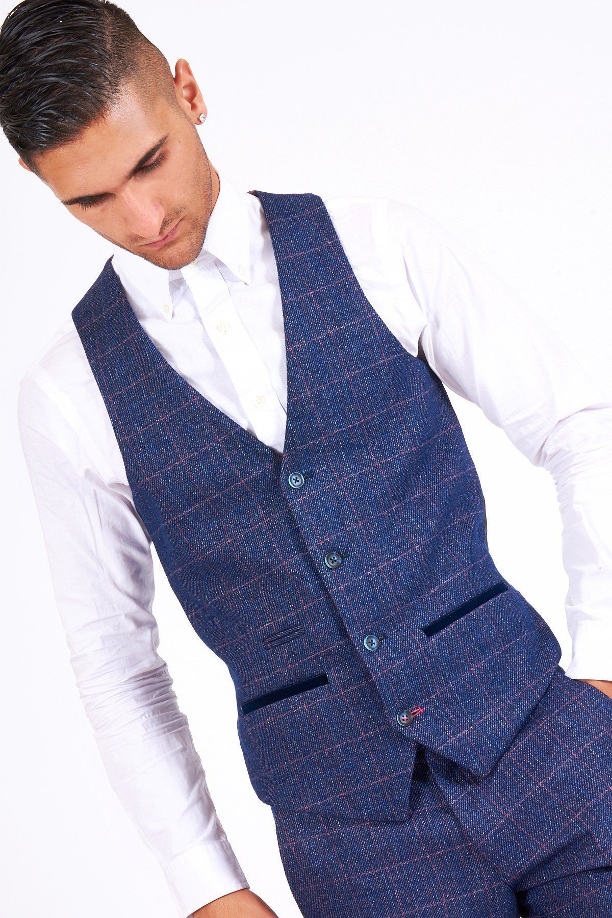 A man poses against a white background in the marcdarcy HARRY Indigo Single Breasted Tweed Check Waistcoat, paired with matching trousers and a white dress shirt, his left hand in his pocket as he looks down.