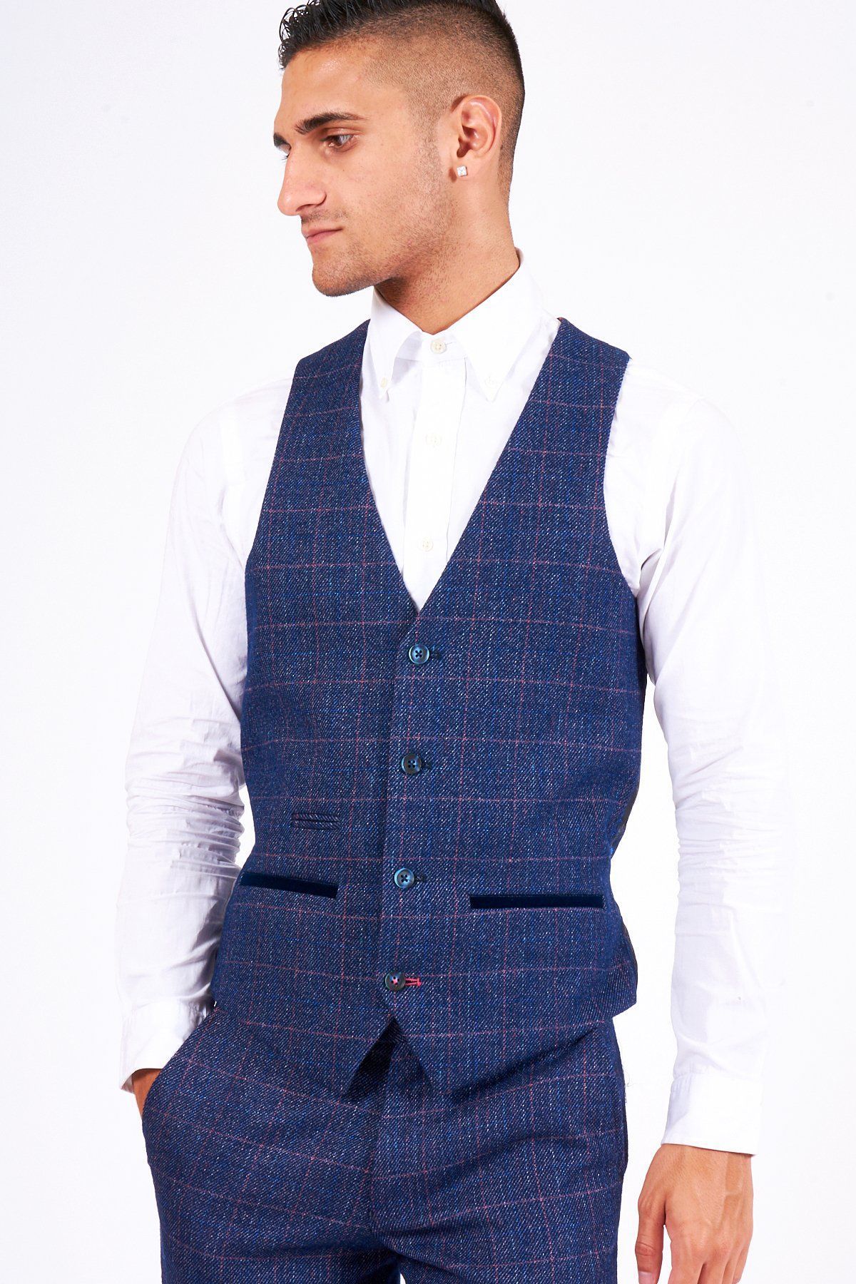 A man wears the marcdarcy HARRY Indigo Single Breasted Tweed Check Waistcoat with matching pants and a white shirt, one hand in his pocket, gazing to the side against a plain white backdrop.