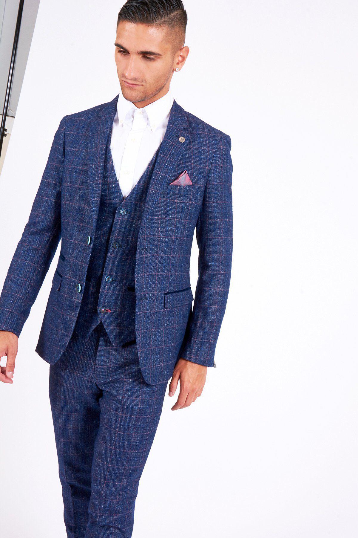A man stands against a plain white background wearing the marcdarcy HARRY Indigo Tweed Check Blazer as part of a blue check three-piece suit, paired with a white shirt and blush pink pocket square, looking down and slightly away.