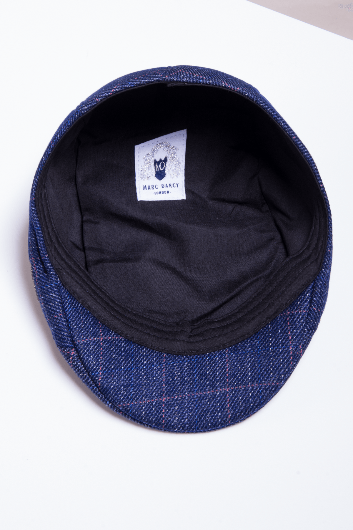 The HARRY - Indigo Tweed Check Flat Cap by Accessory is displayed upside down on a white surface, revealing its black inner lining and a white Marc Darcy London label, highlighting its blue and red check pattern for a vintage look.