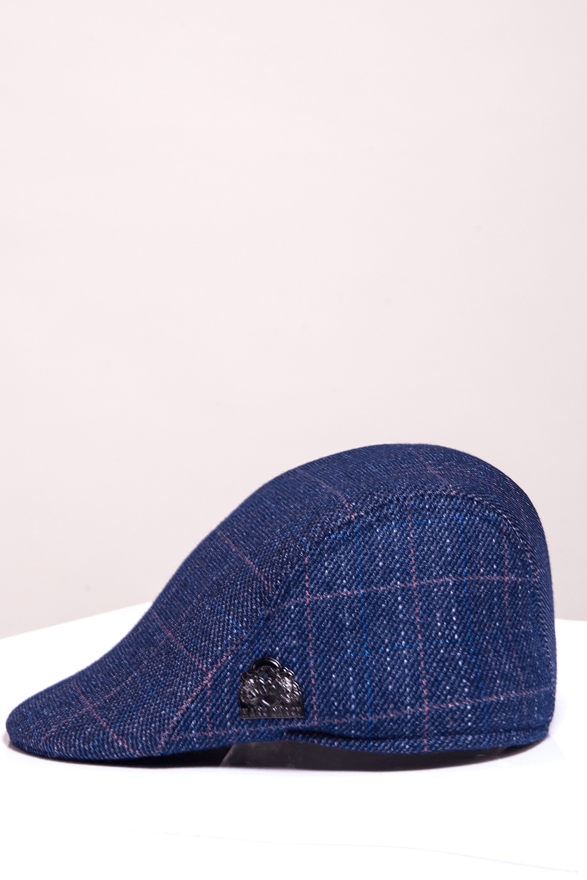 The HARRY - Indigo Tweed Check Flat Cap by Accessory features subtle red checks and a signature badge, shown on a white surface with a light beige background. This stylish cap adds vintage flair to any outfit.