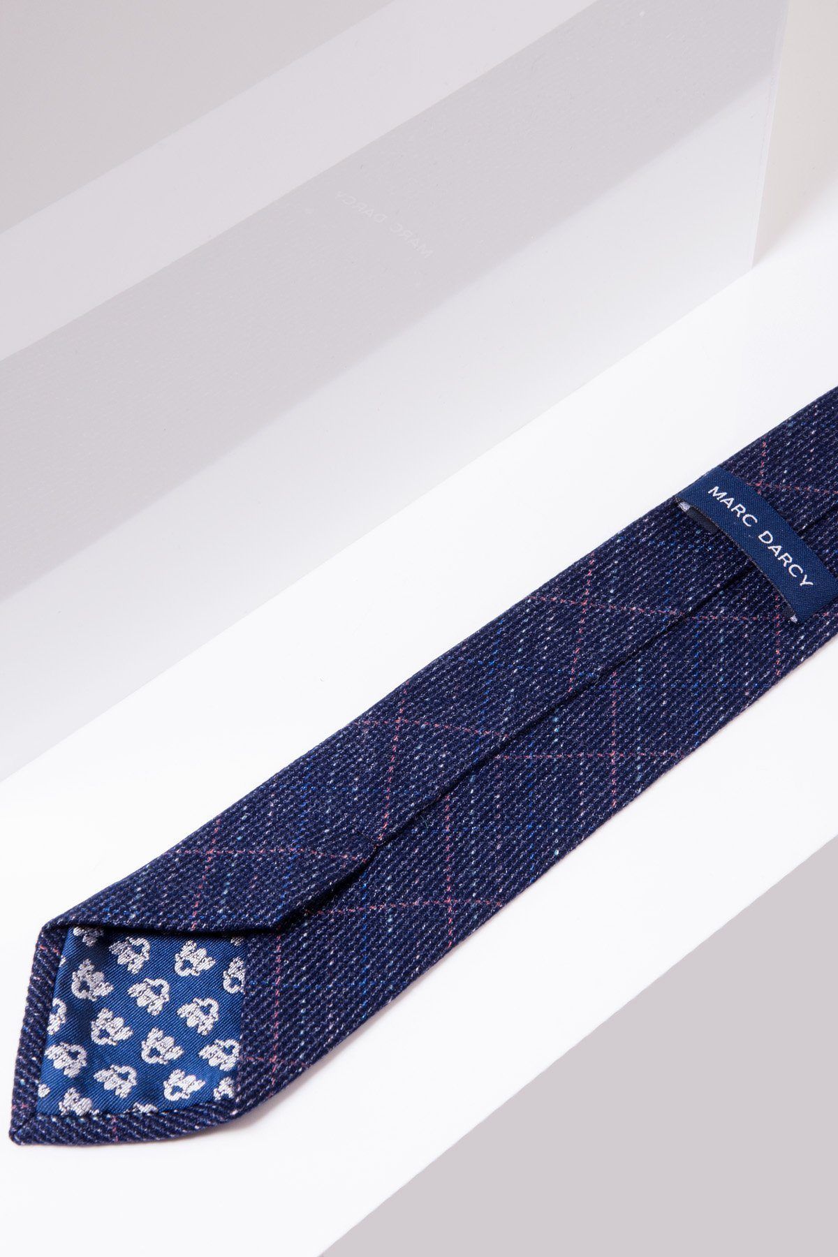 The HARRY Indigo Tweed Check Tie by marcdarcy features a navy textured design with subtle red lines, floral pattern lining, and brand label—ideal for formal occasions and shown on a white and gray background.
