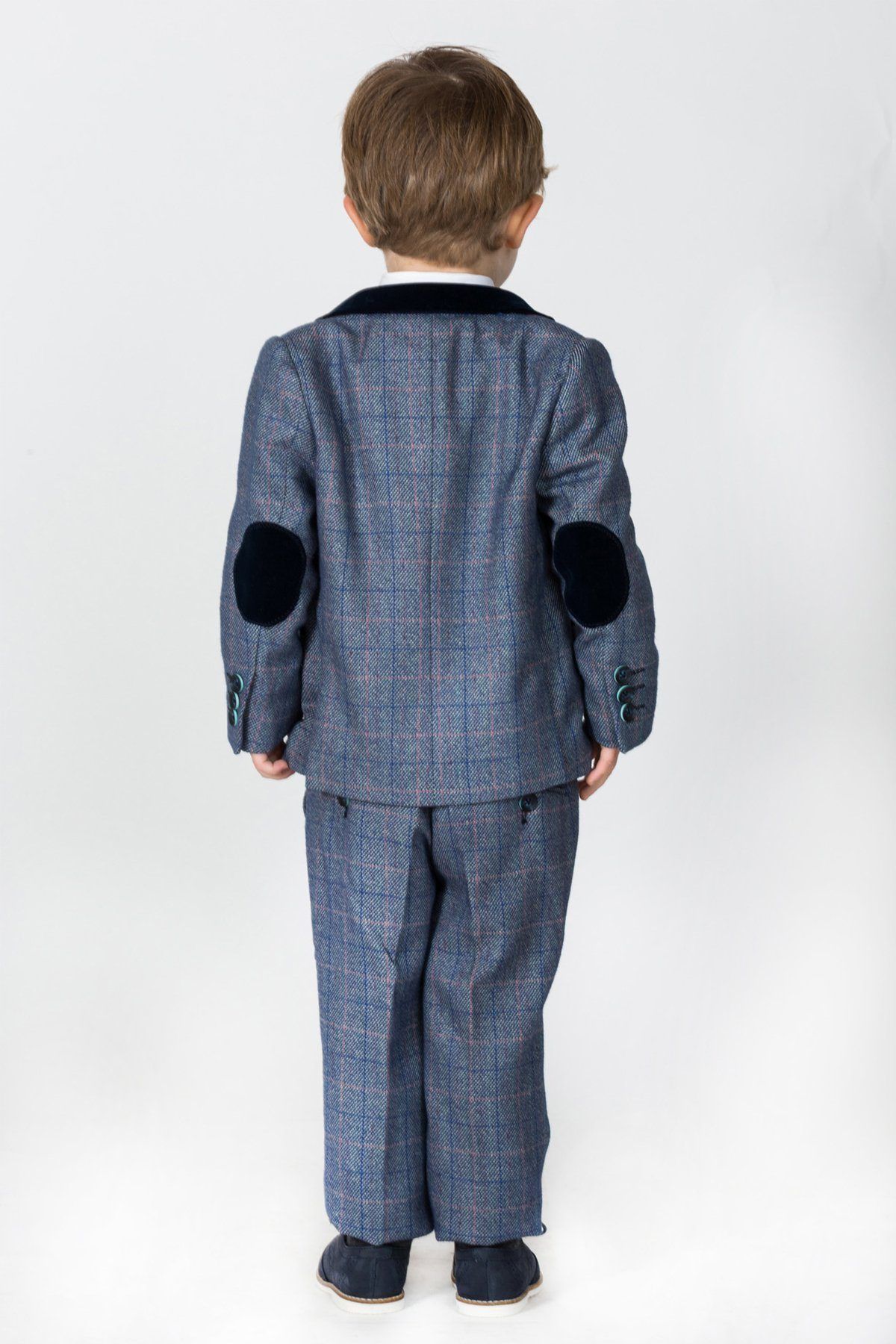 A young boy faces away from the camera in a marcdarcy HILTON Children's Blue Tweed Check Three Piece Suit, featuring dark velvet elbow patches and a matching collar, set against a white background.