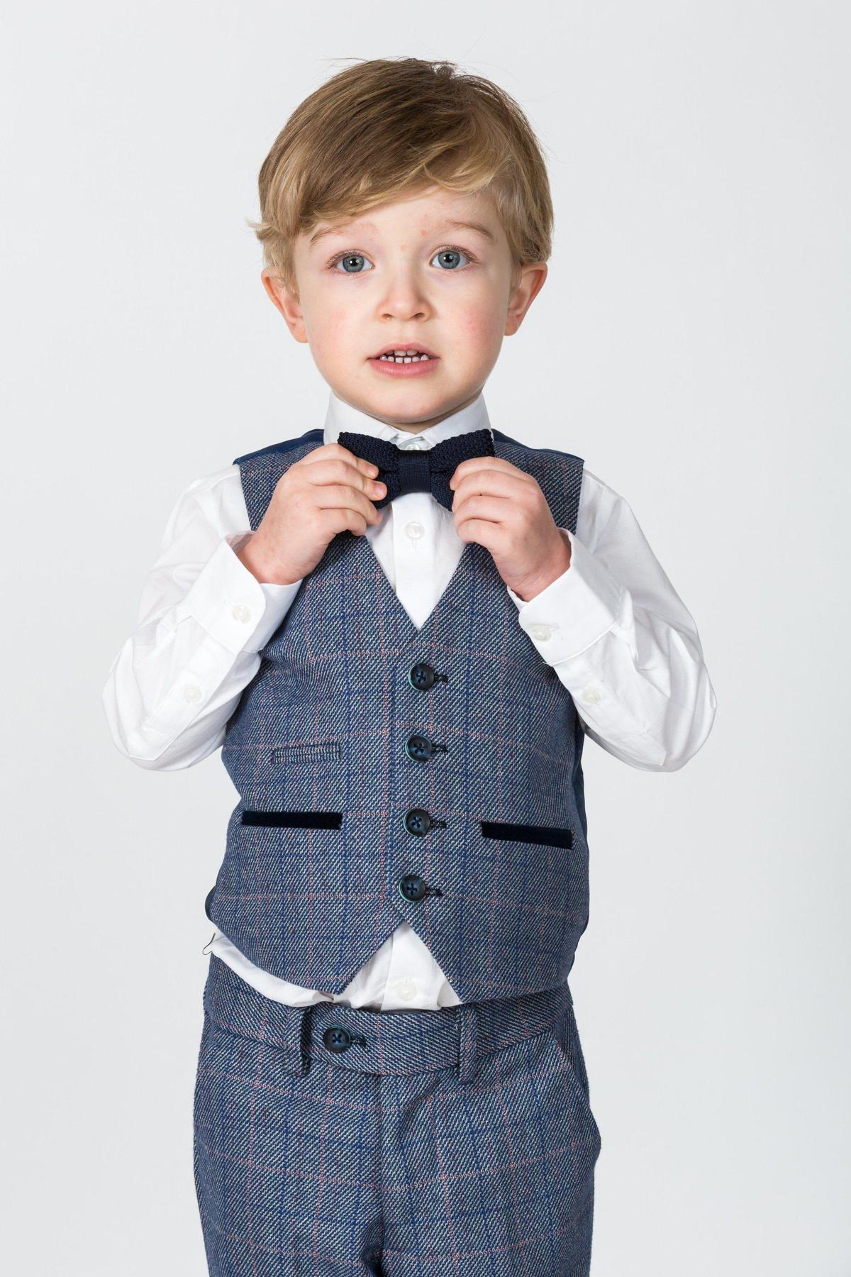 A young boy in the marcdarcy HILTON Childrens Blue Tweed Check Three Piece Suit adjusts his black bow tie over a white shirt, standing against a plain white background.