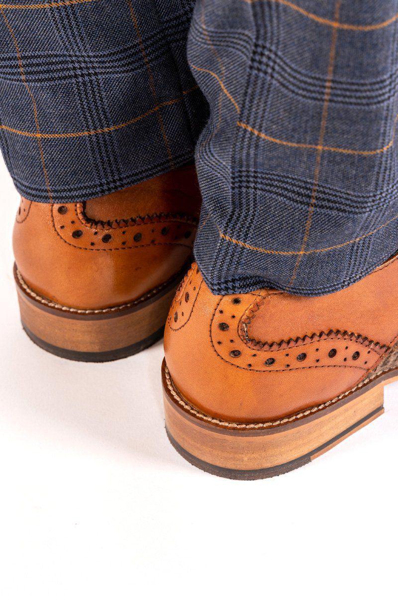 Close-up of a person wearing Marc Darcy JACOB - Tan Leather Tweed Contrast Brogue Shoes with blue plaid trousers featuring a yellow stripe, standing on a white surface. The image highlights the heels and lower legs.
