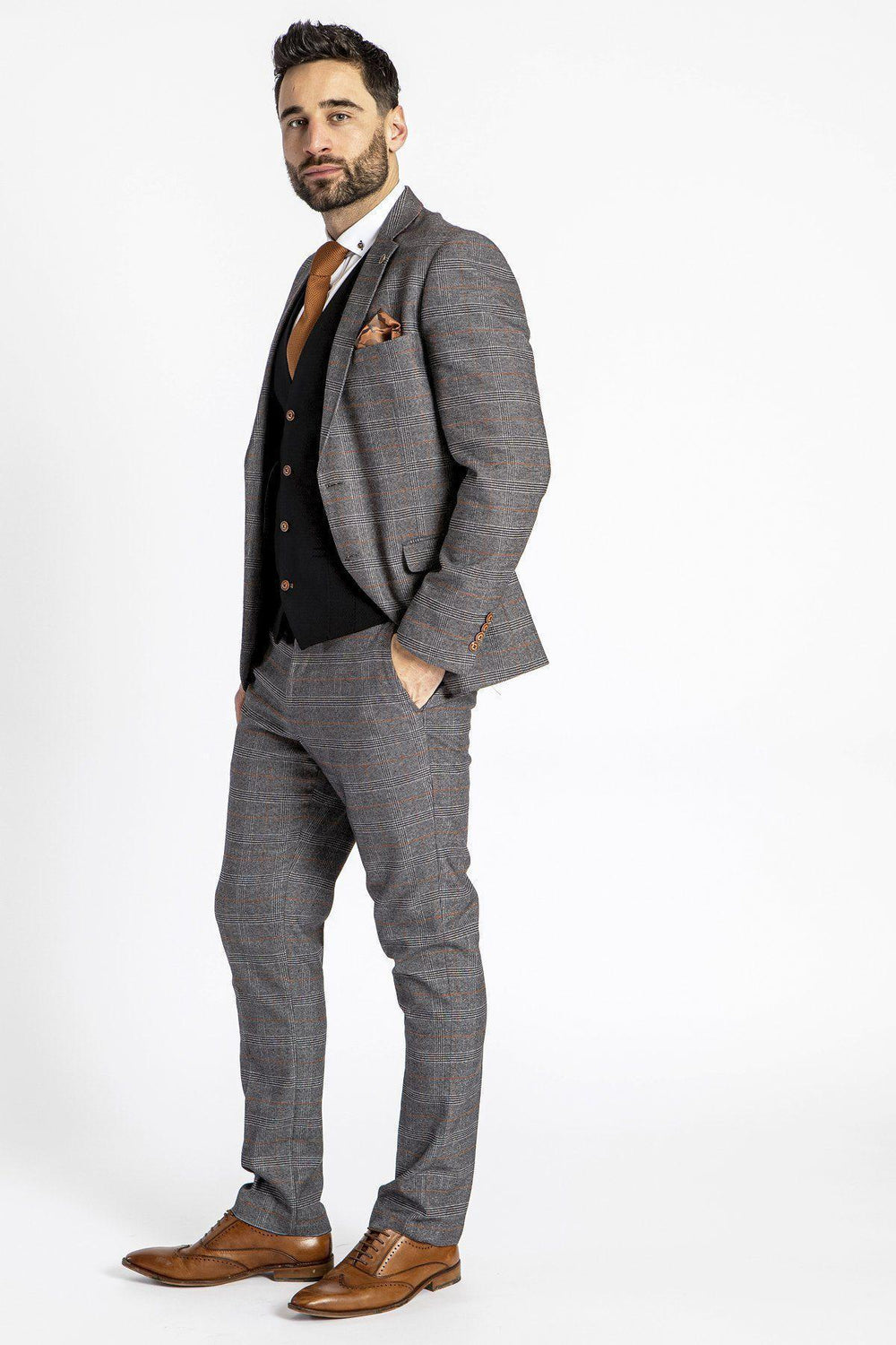 A man with dark hair and a beard stands confidently against a white background, wearing the marcdarcy JENSON Grey Check Suit with MAX Black Waistcoat, paired with an orange tie and brown dress shoes. One hand rests in his pocket.