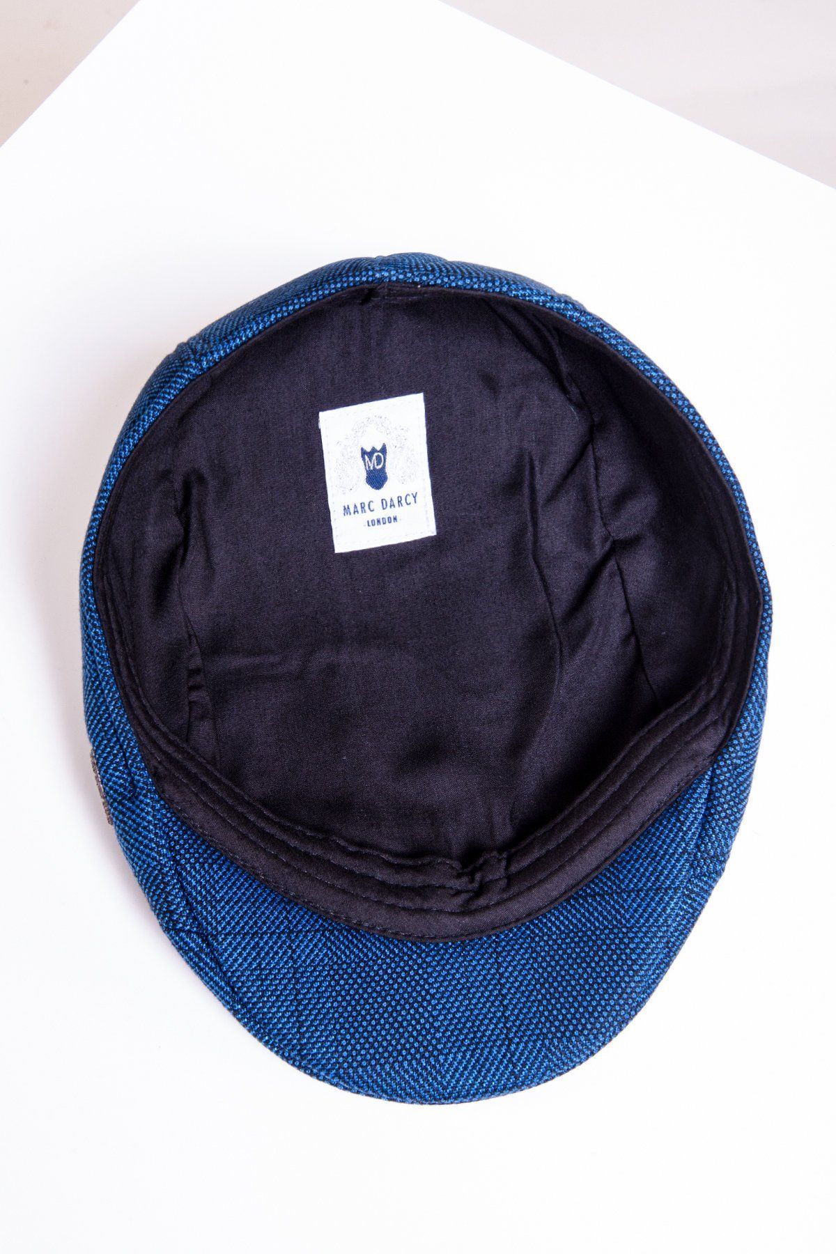 The JERRY - Blue Check Flat Cap by Accessory is displayed inside out, revealing its black inner lining and a white Marc Darcy London label with logo. The blue check cap, exuding vintage style, rests on a white surface.