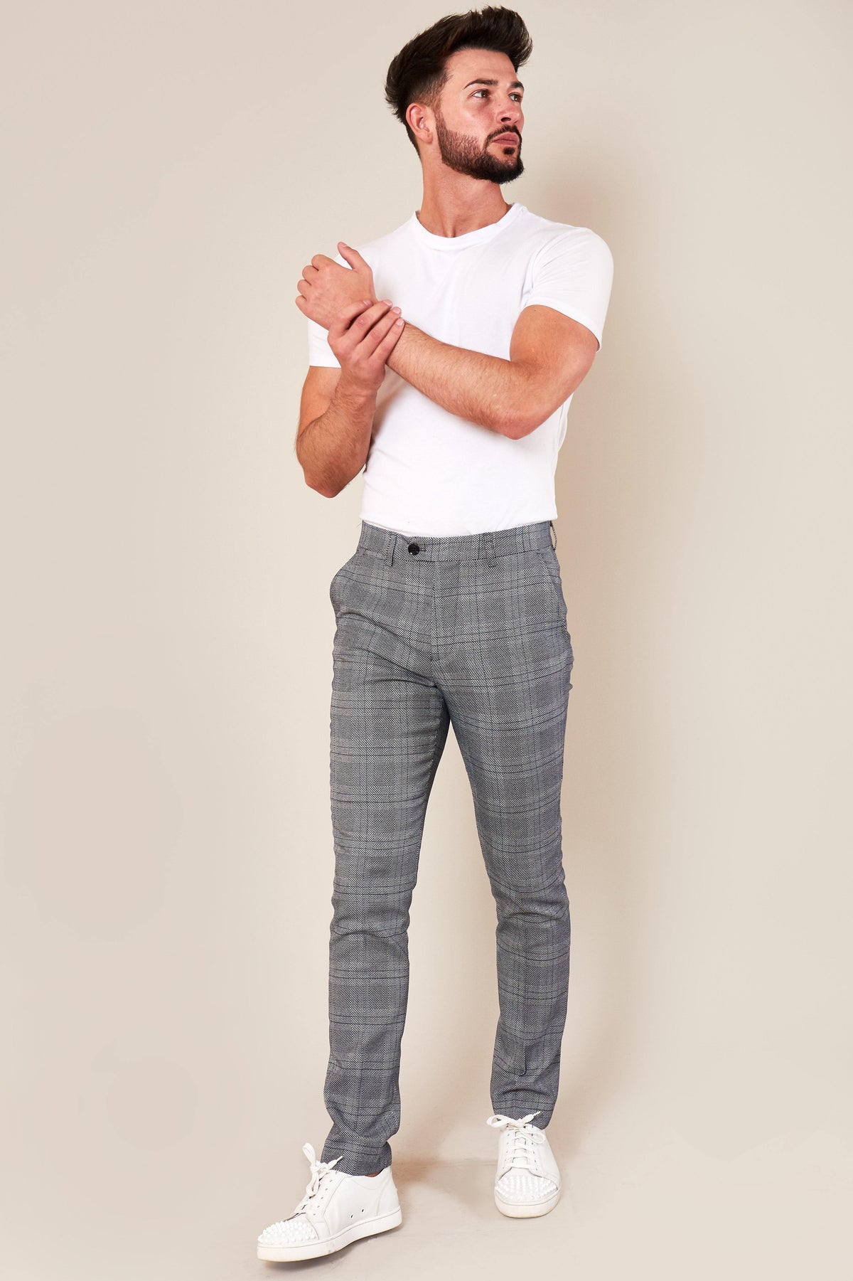 A man with dark hair and a beard wears a fitted white t-shirt, marcdarcy JERRY skinny fit grey check trousers, and white sneakers. He stands against a plain beige background, looking to the side with one arm across his chest.