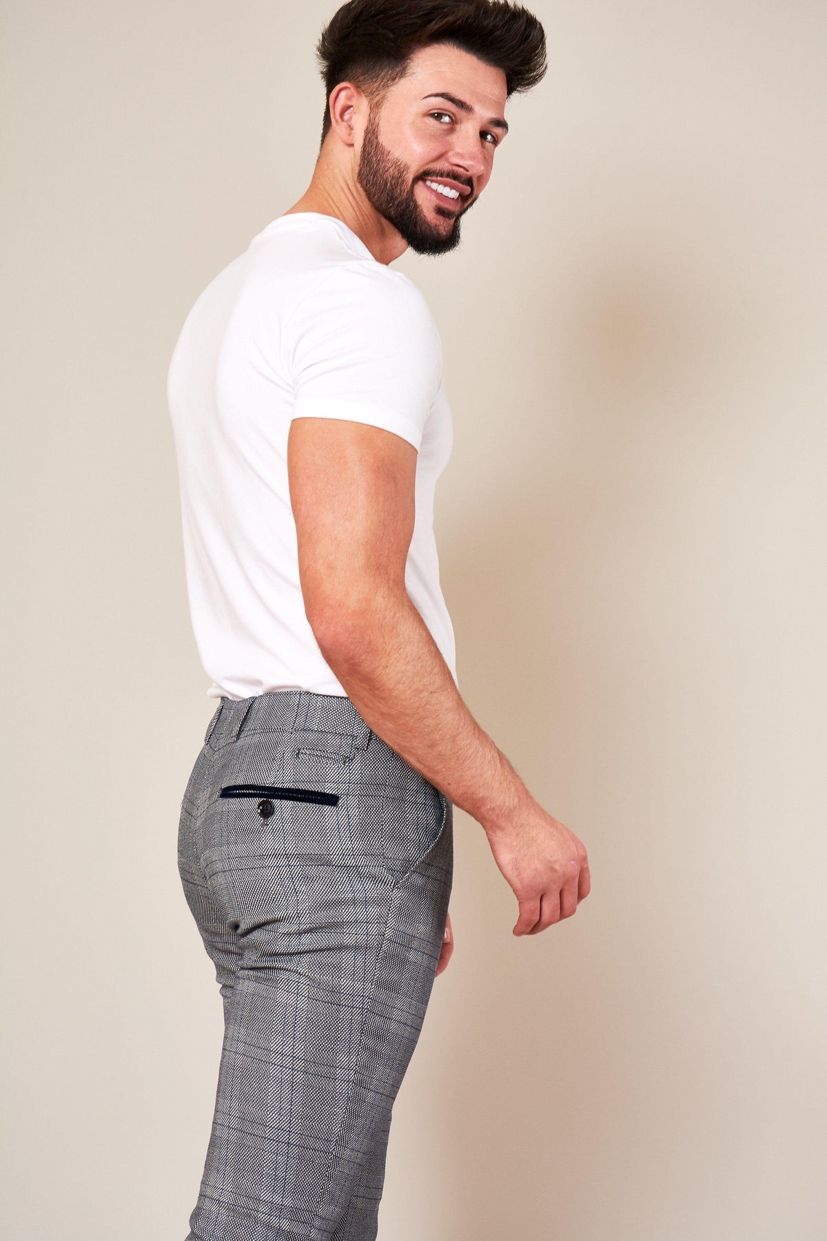 A man with a beard and dark hair smiles over his shoulder, wearing a white t-shirt paired with marcdarcy JERRY Skinny Fit Grey Check Trousers.