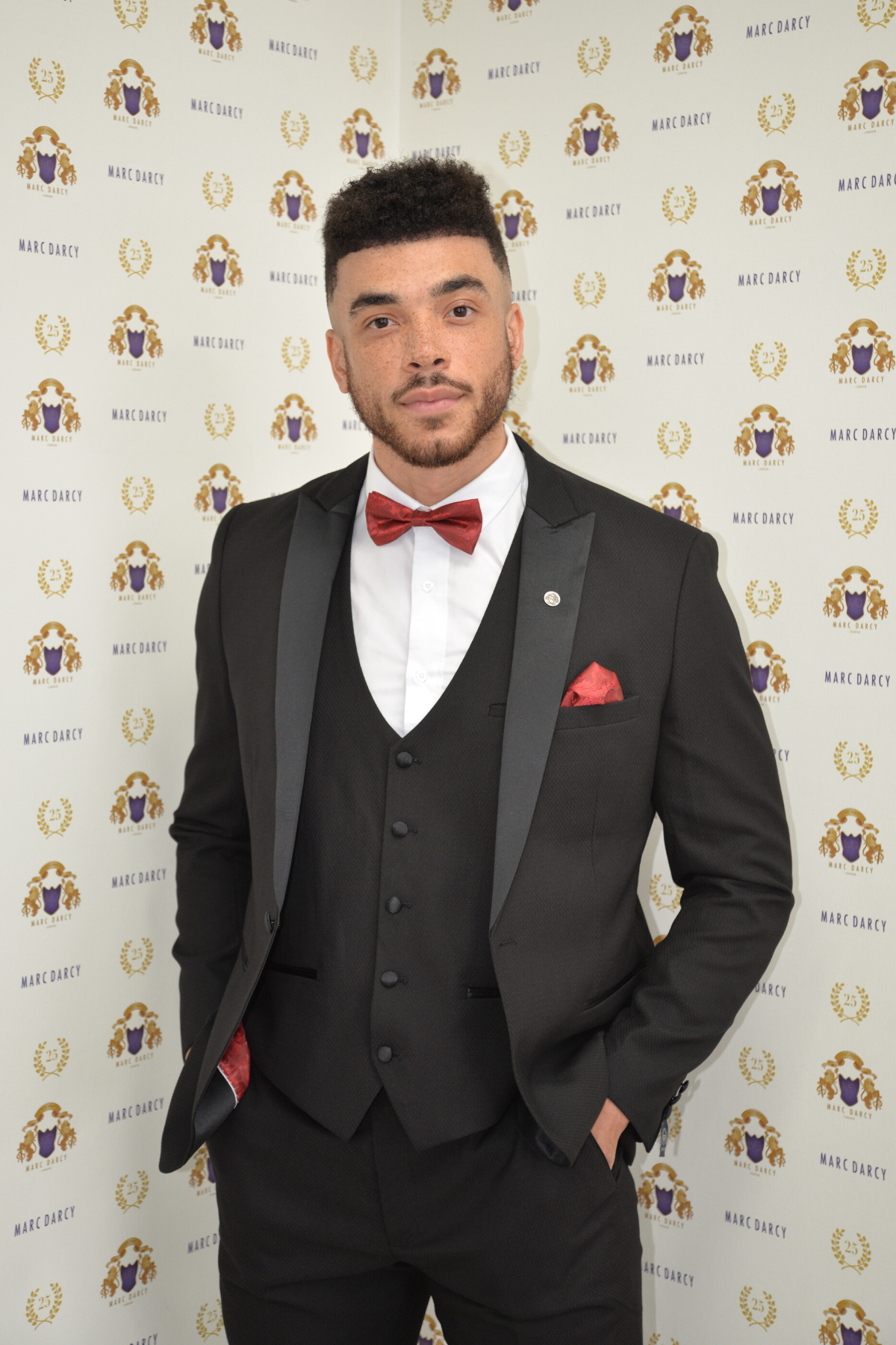 Emmerdale’s Jurell Carter (Nate Robinson) wears the marcdarcy Dalton Black Tux—ideal for black tie events—standing against a marcdarcy-branded backdrop in classic formal style.