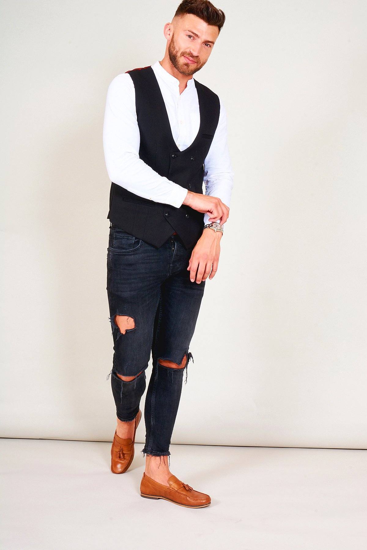 A man in a white shirt, marcdarcy KELLY Black Double Breasted Waistcoat, ripped black jeans, and brown loafers stands against a plain background, adjusting his watch in smart casual style while facing the camera.