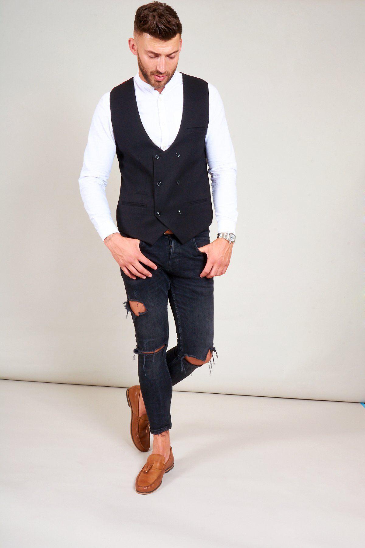 A man in smart casual style wears a white button-up shirt, marcdarcy’s KELLY black double-breasted waistcoat, ripped black jeans, and brown loafers. He stands against a plain background, looking down with one hand in his pocket.