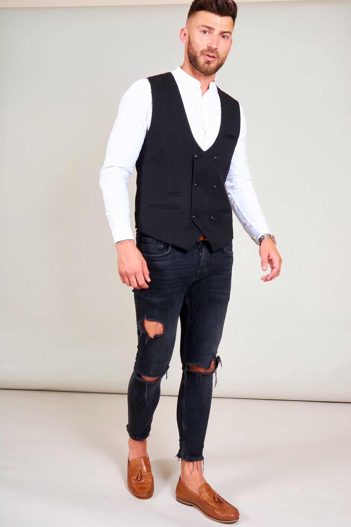 Man wears the marcdarcy KELLY black double breasted waistcoat over a white mandarin collar shirt, paired with distressed black jeans and brown tassel loafers for a smart casual look.
