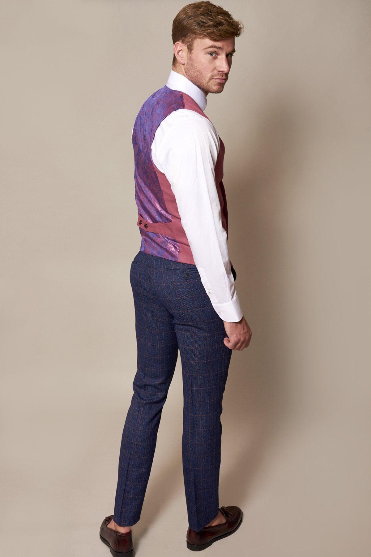 A man, turned slightly away against a beige background, wears a white dress shirt, marcdarcy’s KELLY Pink Double Breasted Waistcoat, navy plaid trousers, and brown shoes while looking over his shoulder.
