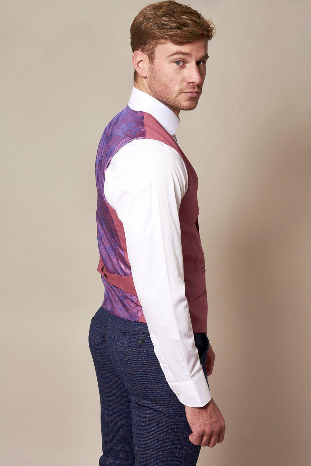 A man with light brown hair and a trimmed beard wears a white shirt, blue checkered trousers, and the marcdarcy KELLY Pink Single Breasted Waistcoat with purple paisley lining. He stands sideways, looking over his shoulder at the camera.