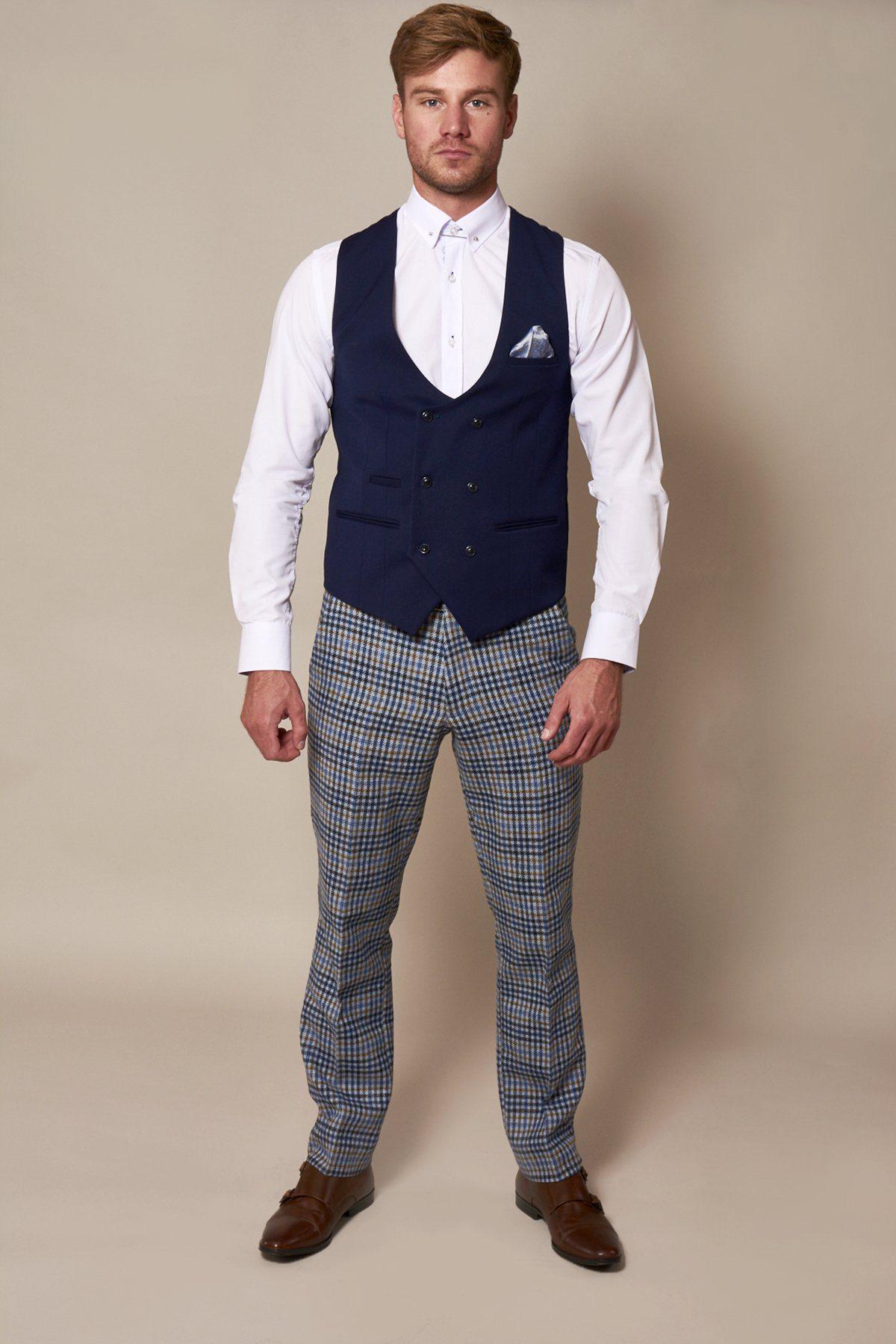 A man with short hair and a beard stands against a plain background, wearing the marcdarcy KELLY Royal Blue Double Breasted Waistcoat, white dress shirt, checked trousers, brown dress shoes, and a pocket square. He faces the camera directly.