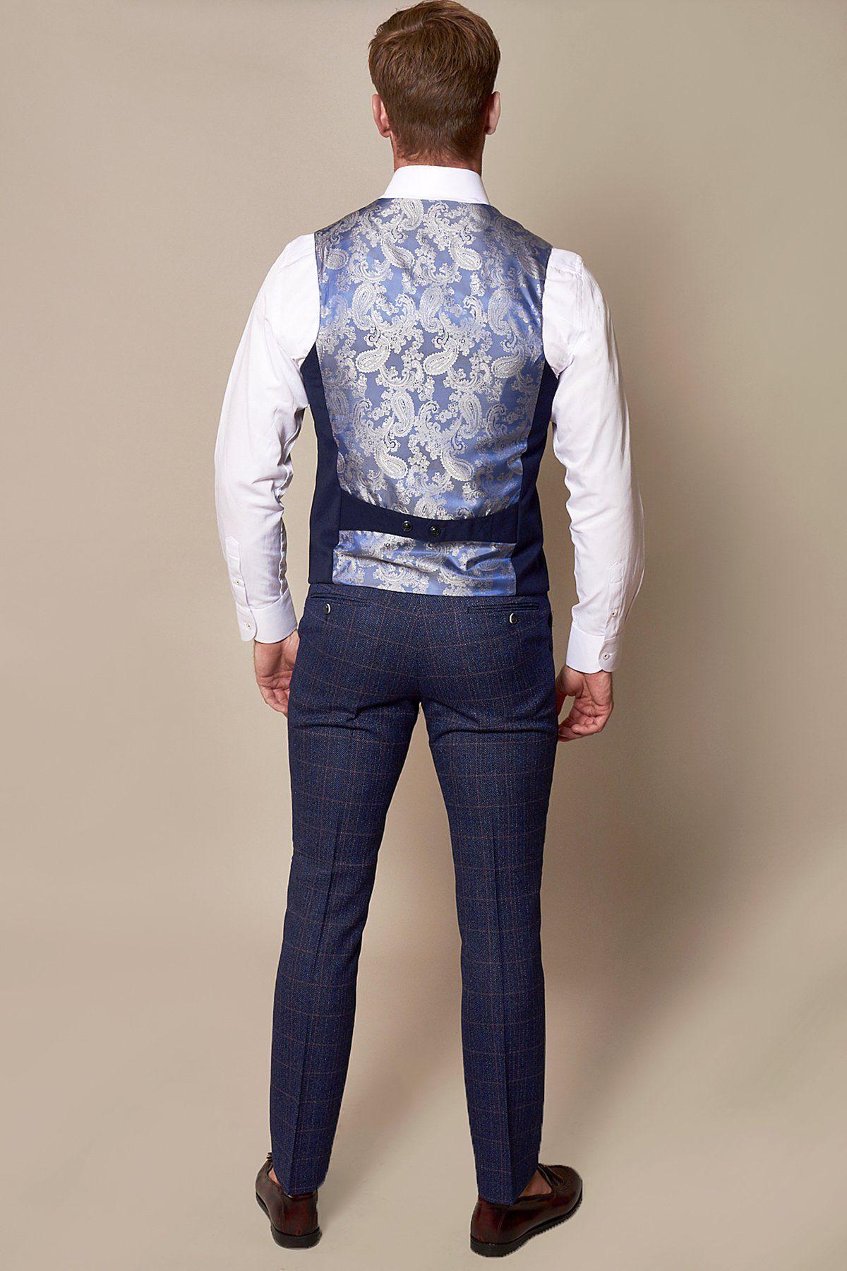 A man stands with his back to the camera, wearing the marcdarcy KELLY Royal Blue Single Breasted Waistcoat, a white dress shirt, blue checked trousers, brown dress shoes, and a shiny light blue floral-back waistcoat against a beige background.