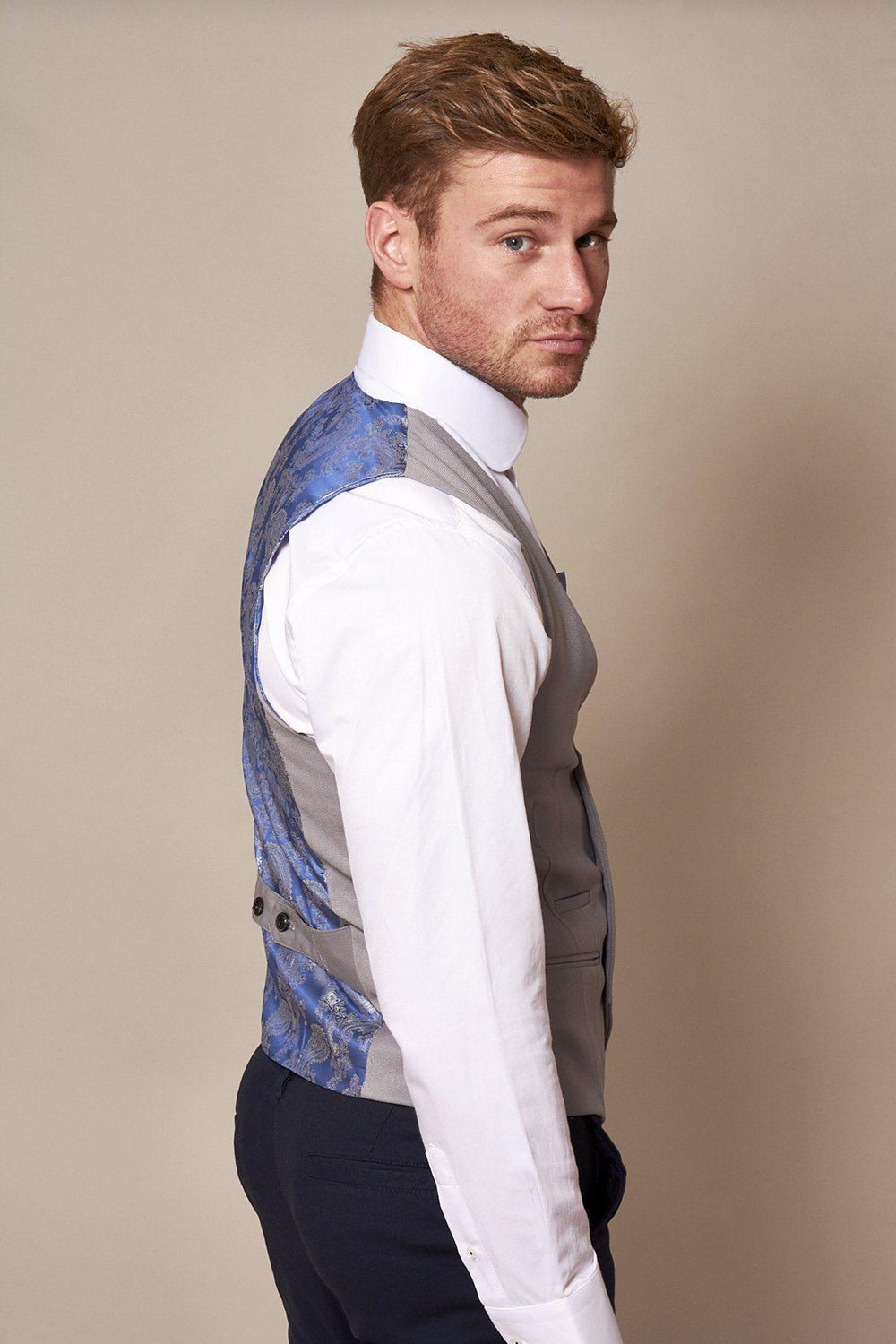 A man with light brown hair and stubble wears the marcdarcy KELLY - Silver Grey Double Breasted Waistcoat, featuring a blue patterned back, styled with a white shirt and blue pants as he looks over his shoulder at the camera.