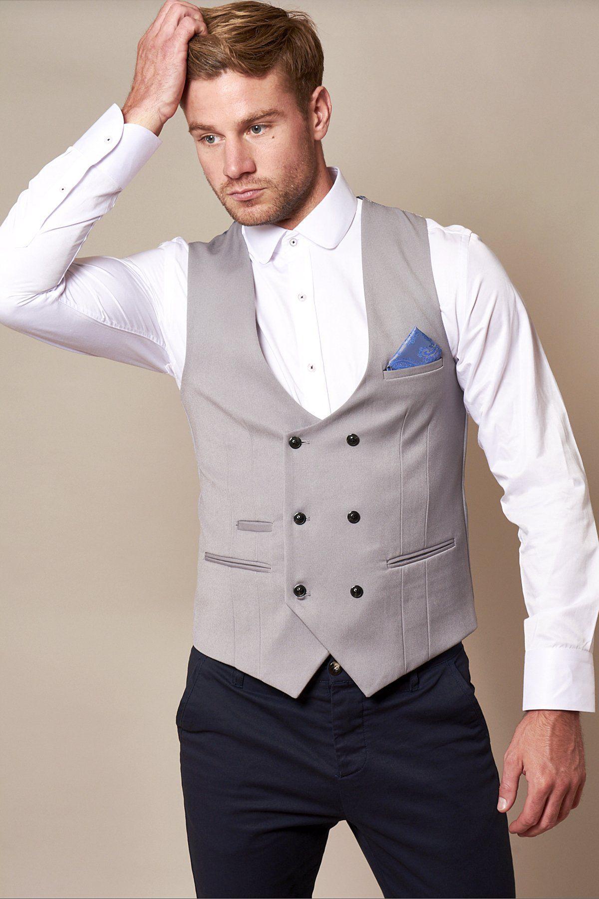 A man wearing the marcdarcy KELLY Silver Grey Double Breasted Waistcoat, white dress shirt, and dark trousers stands against a beige background, touching his hair with one hand.