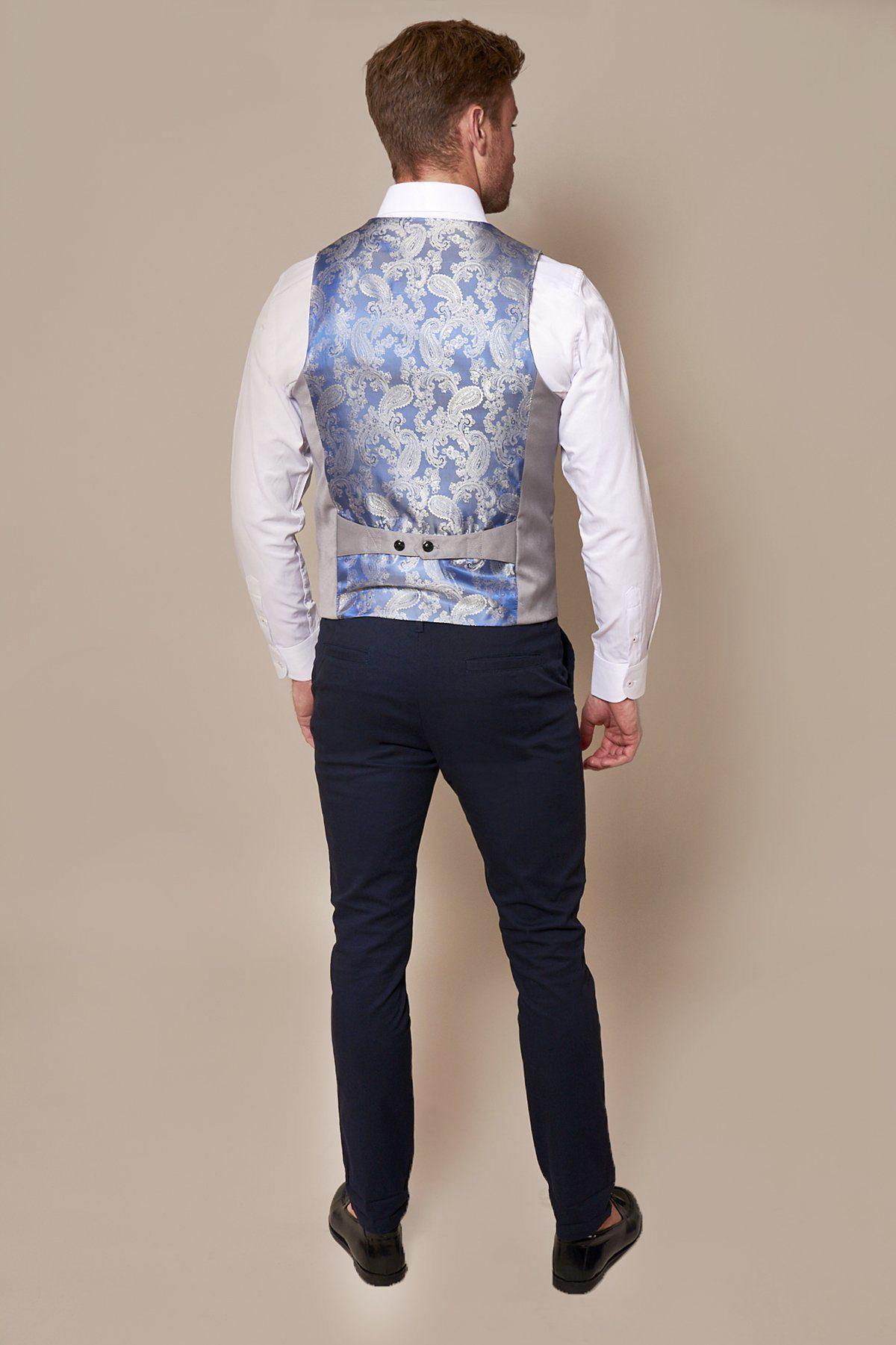 A man faces away in a white shirt, dark pants, black shoes, and the marcdarcy KELLY Silver Grey Double Breasted Waistcoat. The waistcoat features a floral brocade pattern and adjustable strap. Background is plain beige.
